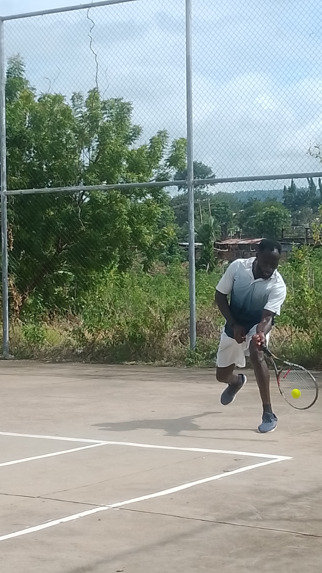 An adult male tennis player is captured in mid-action on an outdoor concrete court, identified as being in Lokoja, Nigeria. The player, of dark complexion, wears a white and blue gradient short-sleeved polo shirt, white shorts, and dark athletic shoes. He is positioned in a dynamic, bent stance, leaning forward and angled to his left, in the process of executing a forehand stroke with a dark-framed tennis racket. A bright yellow tennis ball is visible immediately above the racket strings, indicating contact or imminent impact. A distinct dark shadow of the player and racket is cast on the light grey concrete court surface, which is marked with clear white lines defining sections of the playing area. The court is bordered by a silver chain-link fence supported by metal posts; a thin, dark, winding line, possibly a vine or crack, is visible on the upper left section of the fence. Behind the fence, dense green vegetation consisting of bushes and trees forms the immediate background. In the distant background, beyond the foliage, multiple structures and undulating hills are discernible beneath an overcast, light grey sky.