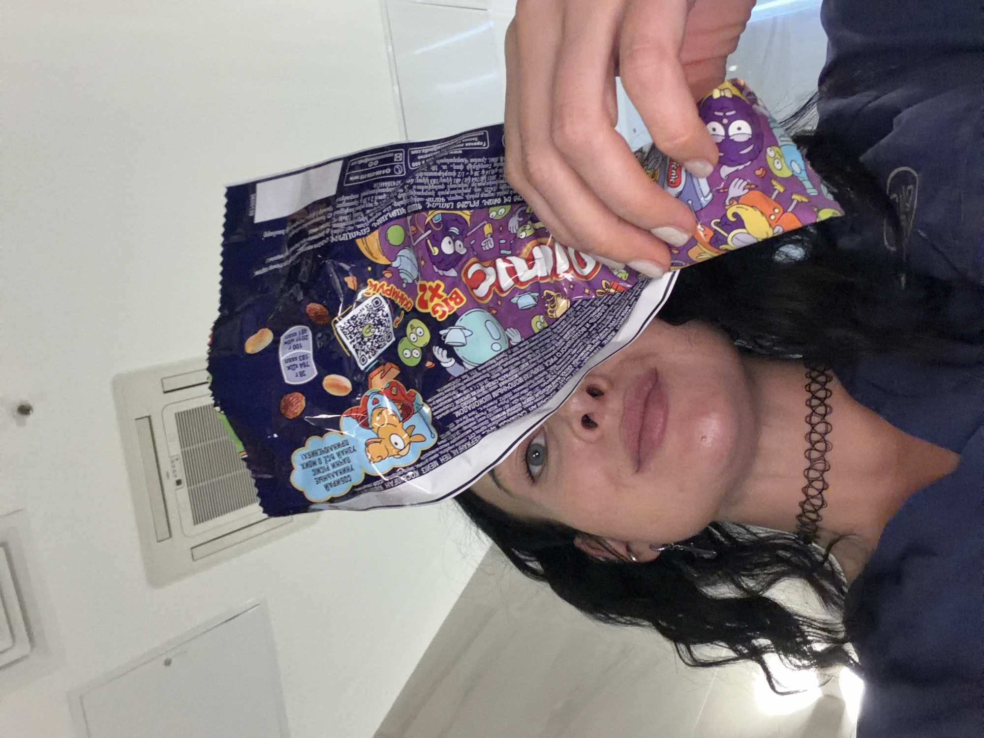 A woman with dark hair, a choker, and an earring is holding a bag of "Big Crunch" snacks in front of her face. The bag is purple and has cartoon characters on it. The text on the bag is in Russian and includes phrases like "УЗНАЙ ВСЁ О МОИХ ПРИКЛЮЧЕНИЯХ!". She is indoors, possibly in a bathroom, as there is a white wall and a ventilation unit visible in the background.