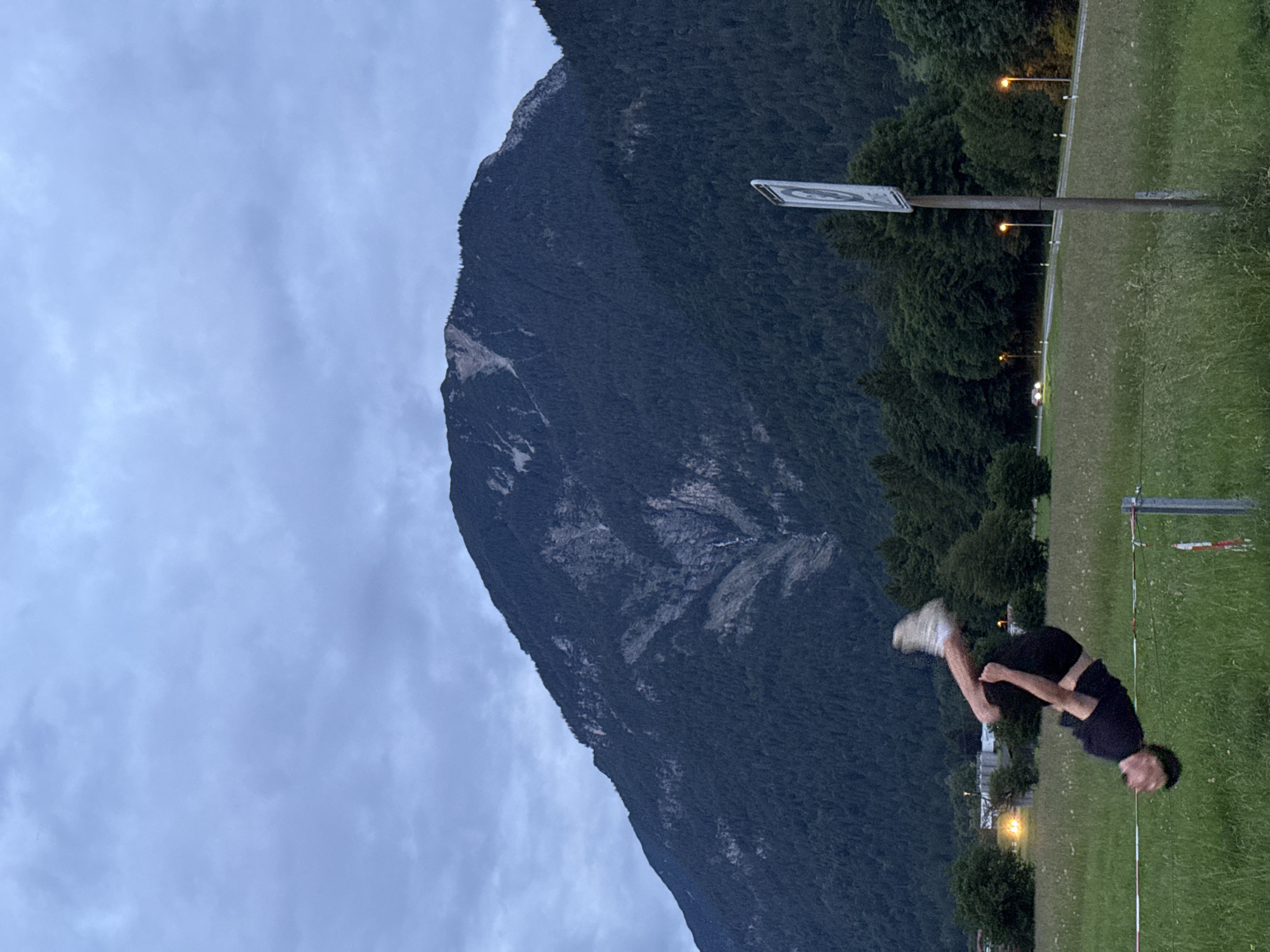 The image depicts an outdoor evening scene in Garmisch-Partenkirchen, Germany, with the Wank mountain dominating the background. A male individual is captured mid-air, performing a tucked backflip over a red and white striped tape fence in a grassy foreground.
The central subject, a male, is oriented horizontally, with his head positioned towards the lower left and his legs tucked towards his chest. He wears a dark (likely black) t-shirt, dark shorts, and light-colored sneakers, exhibiting motion blur on his extremities. He is suspended above a two-strand fence made of white and red striped tape, supported by metal posts on the right. The fence stretches across the lower portion of a green, unkempt grassy field.
In the mid-ground, a paved road curves from the left towards the right, flanked by several luminous yellow-orange streetlights. A single vehicle with illuminated headlights is visible on the road in the distant center. A dense forest line of coniferous and deciduous trees separates the road from the mountain's base.
The background is dominated by the Wank mountain, a large, dark, and heavily forested peak characteristic of the Bavarian Alps. Its slopes feature patches of exposed grey rock and a prominent vertical lighter streak, indicative of a gully or rockfall, descending near the center of the mountain face. The sky above is entirely overcast with grey clouds, providing a soft, diffused light consistent with dusk or early night, further evidenced by the activated streetlights and vehicle headlights. The overall impression is one of athletic activity set against a dramatic natural and semi-urban landscape on the outskirts of Garmisch-Partenkirchen.