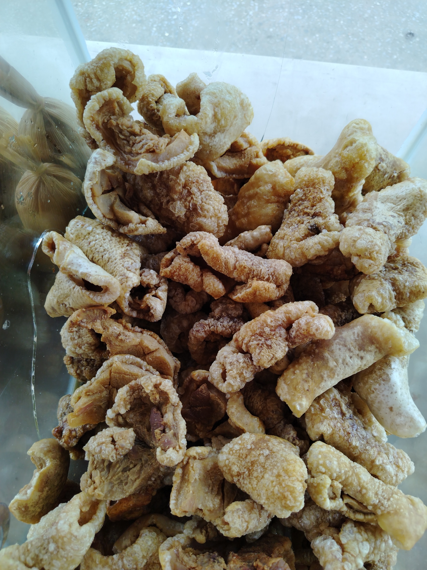 A close-up, high-angle shot shows a clear container filled with crispy, golden-brown chicharrón (pork rinds). The chicharrón pieces are irregular in shape and size, filling the frame. At the edge of the container, wrapped bundles are visible. The location is Tinaquillo, Venezuela.