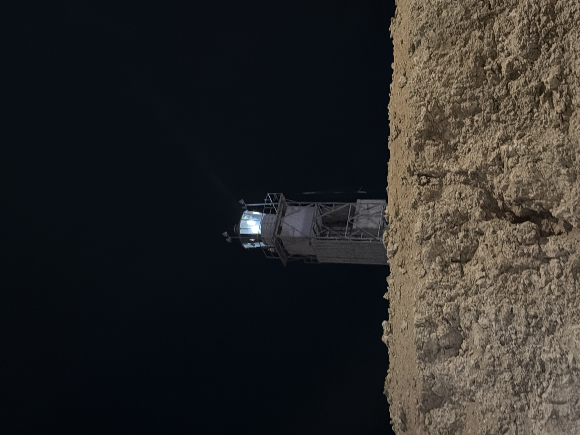 The image depicts a lighthouse at night, situated atop a rocky, elevated terrain. The lighthouse itself is a tall, slender structure, appearing to be under construction or renovation, with scaffolding visible around its upper portion. The lantern room at the top is illuminated with bright, blue-white light, casting a beam into the dark sky. The foreground consists of rough, textured rock formations, likely part of the landscape of Sharm El Sheikh. The overall atmosphere is one of isolation and the functional presence of the lighthouse against the backdrop of the night.