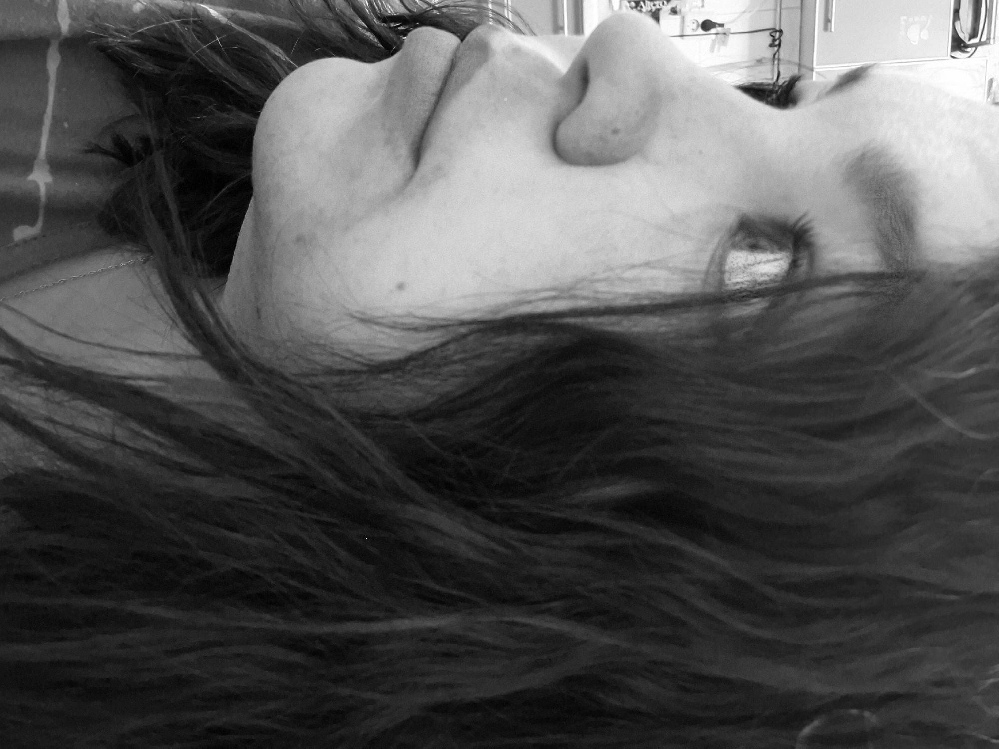 A black and white, low-angle, close-up shot depicts a person with dark hair lying down, facing upwards. The focus is on the face, with the nose and lips prominently visible. The person's eye is partially obscured by hair. In the background, out of focus, is a blurred image of what appears to be a medical environment, potentially a hospital room. There is no explicit indication of the city of Марковский, Russia, present in the image.