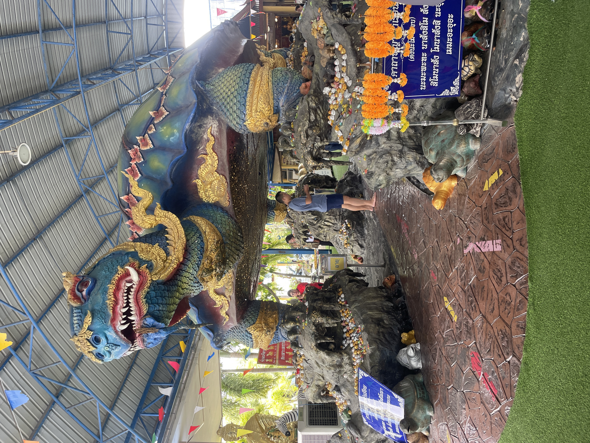 The image shows a large, colorful statue of a mythical turtle, possibly a depiction of a naga or a dragon-like creature, at a temple or shrine in Bang Saray, Thailand. The turtle is blue and green with gold accents and a fierce facial expression. It is elevated on a rocky, artificial base adorned with many smaller turtle figurines and offerings. Several people are present, some observing the statue, others seemingly engaged in prayer or making offerings. The environment appears to be an outdoor covered area with a metal roof and supporting structure. Decorative flags and garlands are visible, adding to the festive or religious atmosphere. In the foreground, there are more turtle statues and a blue sign with Thai text. Artificial grass covers a portion of the ground.