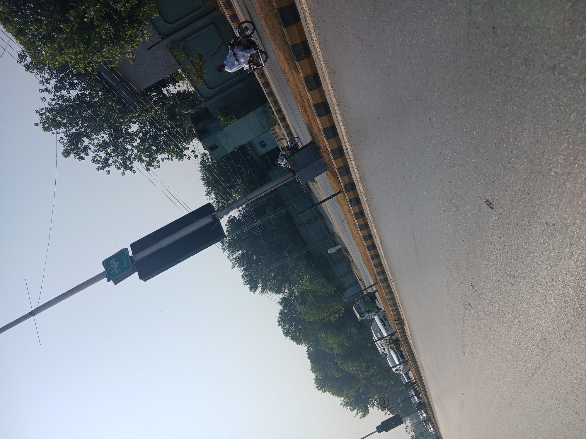 The image depicts a street scene in Peshawar City Tehsil, Pakistan, under a bright sky. A tall street sign with Arabic lettering, appearing to be a street name sign, is prominently visible, angled towards the sky. The sign is green with white text and is attached to a pole. Below this sign is a larger, dark rectangular sign, also angled. The street itself is paved and appears to be a busy road with traffic. Several vehicles are present, including cars, a green auto-rickshaw, and at least two motorcycles. One motorcycle rider, dressed in traditional white clothing, is clearly visible. The road is bordered by a curb with alternating black and yellow markings. Lush green trees line the side of the road, and a building with a green facade and patterned walls is visible in the background. The overall impression is that of an urban or suburban street in Pakistan.