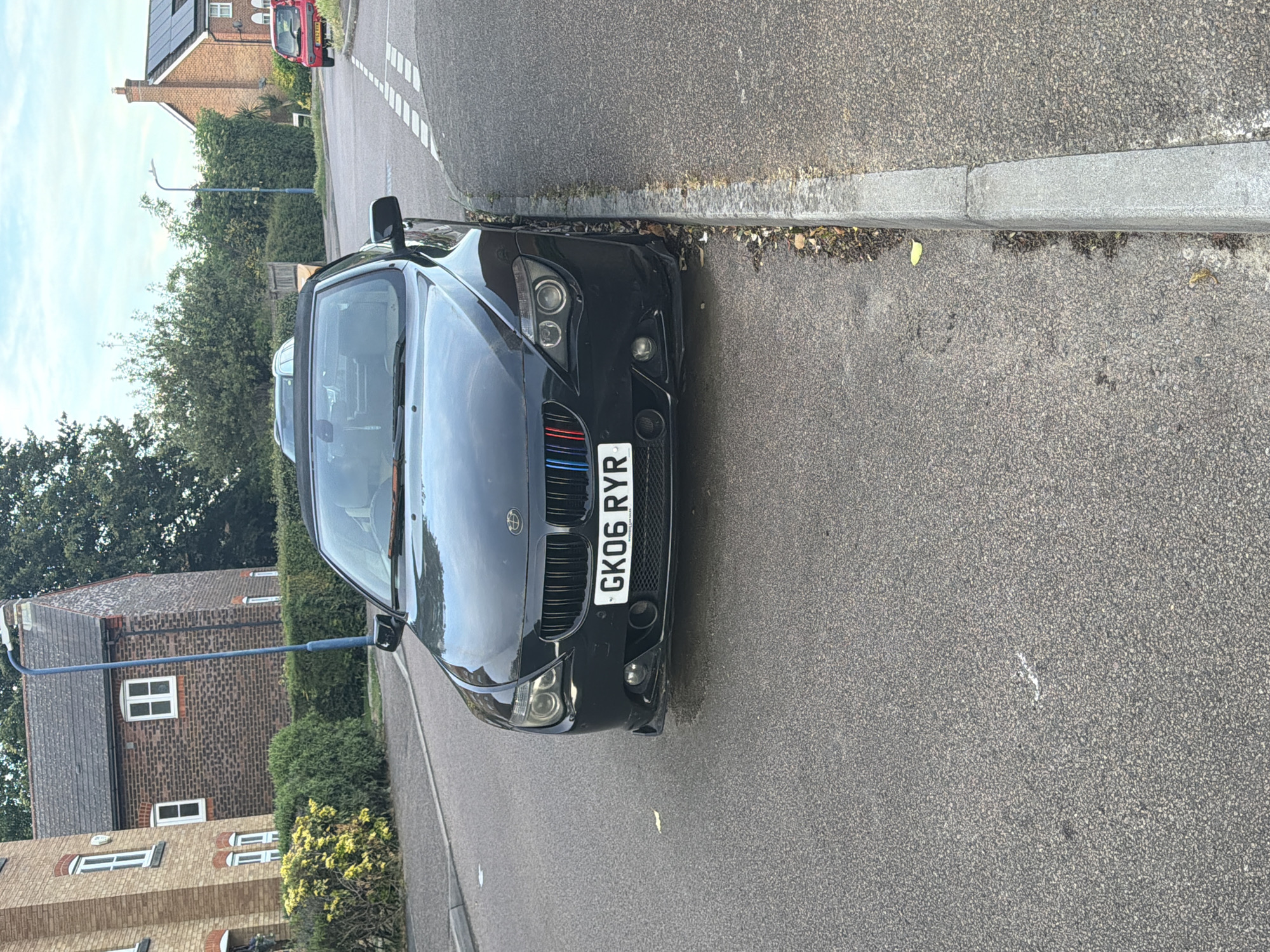 The image displays an eye-level street view of a residential area, likely located in Maidstone, United Kingdom, as indicated by the "GK06 RYR" license plate on the foreground vehicle, which signifies initial registration in the Maidstone or Canterbury area.
In the immediate foreground, a black BMW E63/E64 6 Series convertible is parked parallel to a concrete curb on the left side of an asphalt road. The car's front is angled slightly towards the right side of the frame. Its UK registration plate, "GK06 RYR," is centrally mounted. The left side of the vehicle's kidney grille features decorative red, light blue, and blue vertical stripes. The lower portion of the front bumper on the driver's side exhibits minor localized damage, visible below the left headlight assembly. The driver's side front tire is positioned directly adjacent to the concrete curb. The convertible roof is retracted.
The asphalt road surface appears dry and well-maintained, featuring granular texture. Further down the road on the right, white dashed road markings are visible, indicating an upcoming junction or hazard. A concrete curb separates the road from a narrow verge containing sparse dry leaves and minimal vegetation.
In the midground, beyond the BMW, the street continues with additional parked vehicles and residential properties. On the left, a low hedge with yellow-green foliage is situated behind the parked BMW. Further back on the left, a two-story brick house with white-framed windows and a dark tiled roof is partially visible. A tall, slender, blue-grey street light pole stands in front of this house. On the right side of the street, a red hatchback car, partially identified by the license plate "AY62 ###", is parked facing away from the viewer. Behind this red vehicle, another two-story brick house with a dark tiled roof featuring solar panels is visible. The street is lined with mature green trees and hedges, characteristic of a suburban environment.
The background consists of a partially cloudy blue sky above the treetops and houses, suggesting daylight conditions, possibly late afternoon or early morning, due to the soft, diffused light. No individuals are visible in the scene.