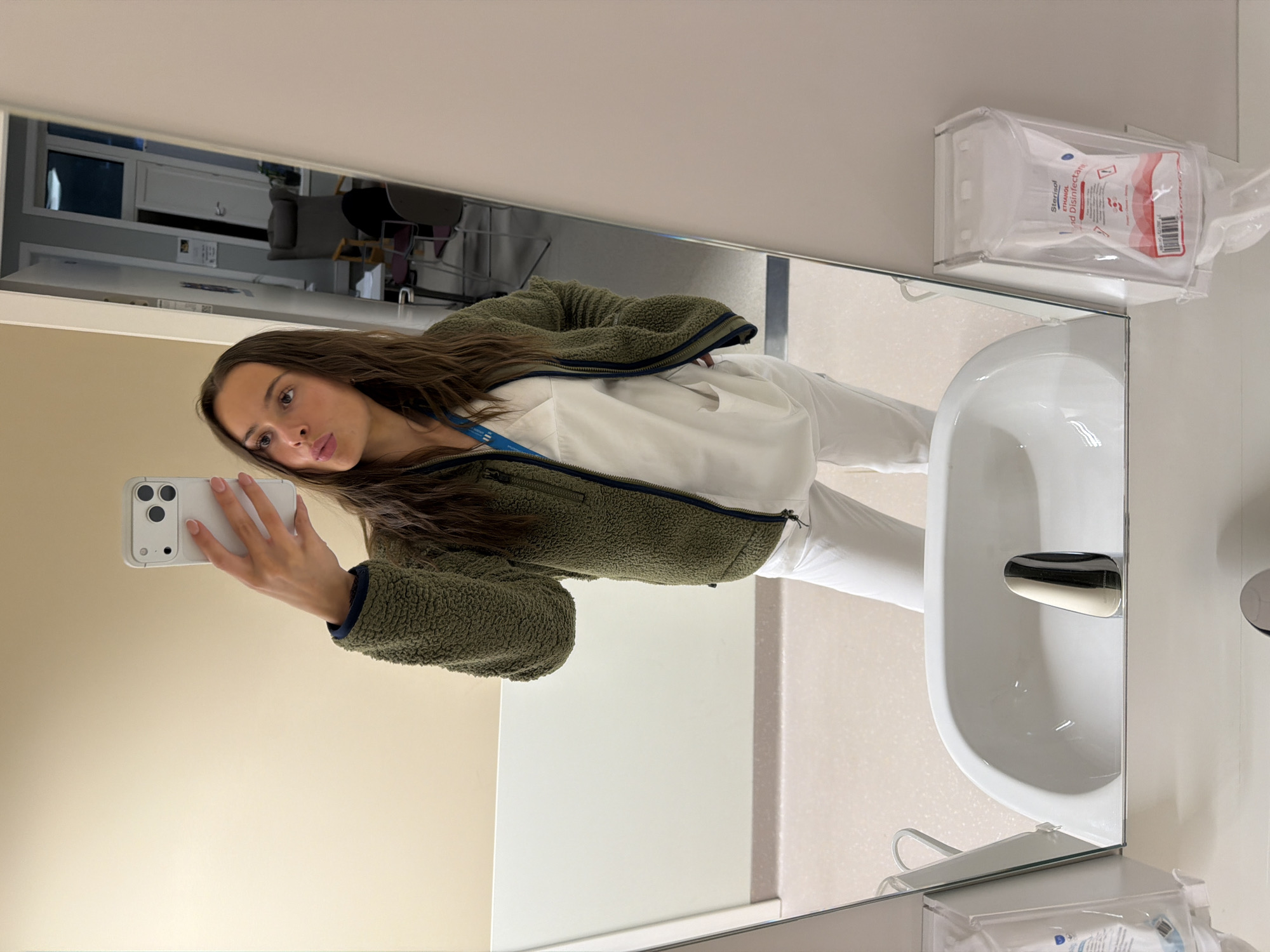 In an indoor setting, possibly a hospital or clinic bathroom in Harstad, Norway, a young woman stands in front of a mirror, taking a selfie. She is wearing a green fleece jacket over a white scrub top and white pants. Her hair is long and brown, and she is making a kissing face at the camera. The mirror reflects a sink with a chrome faucet and a hand sanitizer dispenser on the right. In the background, visible in the mirror's reflection, is a chair, a door, and a window. The overall environment appears clean and well-lit.