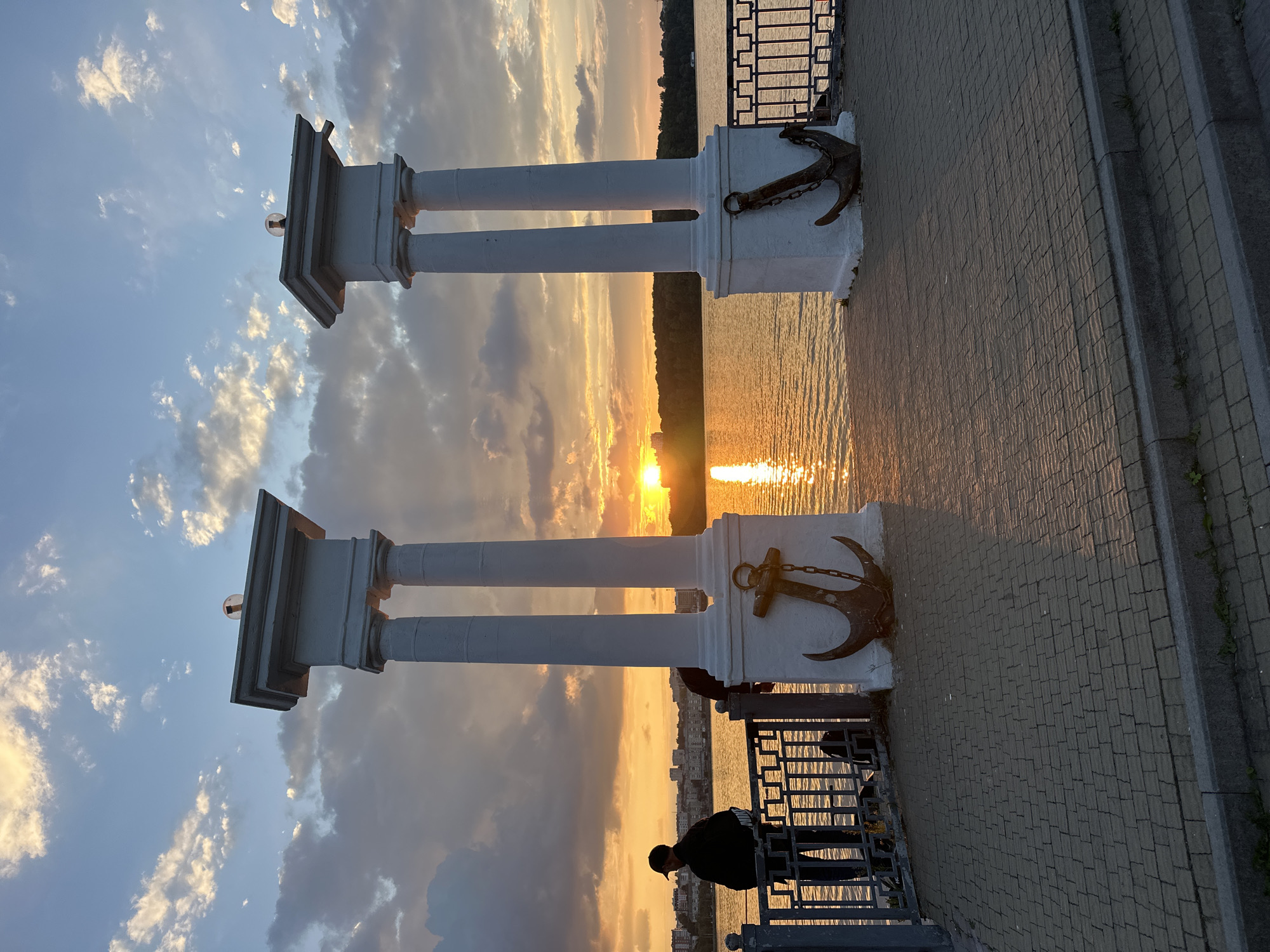 The image shows a sunset over Ternopil Lake, near the Ternopil Castle. Two white columns stand on the embankment, each topped with a lamp. Anchors are displayed at the base of the columns. A person in a black jacket and cap stands by a railing, looking out at the lake. The sun is setting on the horizon, casting a golden glow on the water and the surrounding buildings. The sky is filled with clouds, some of which are illuminated by the setting sun. The embankment is paved with cobblestones, and there is a metal fence along the edge of the water.