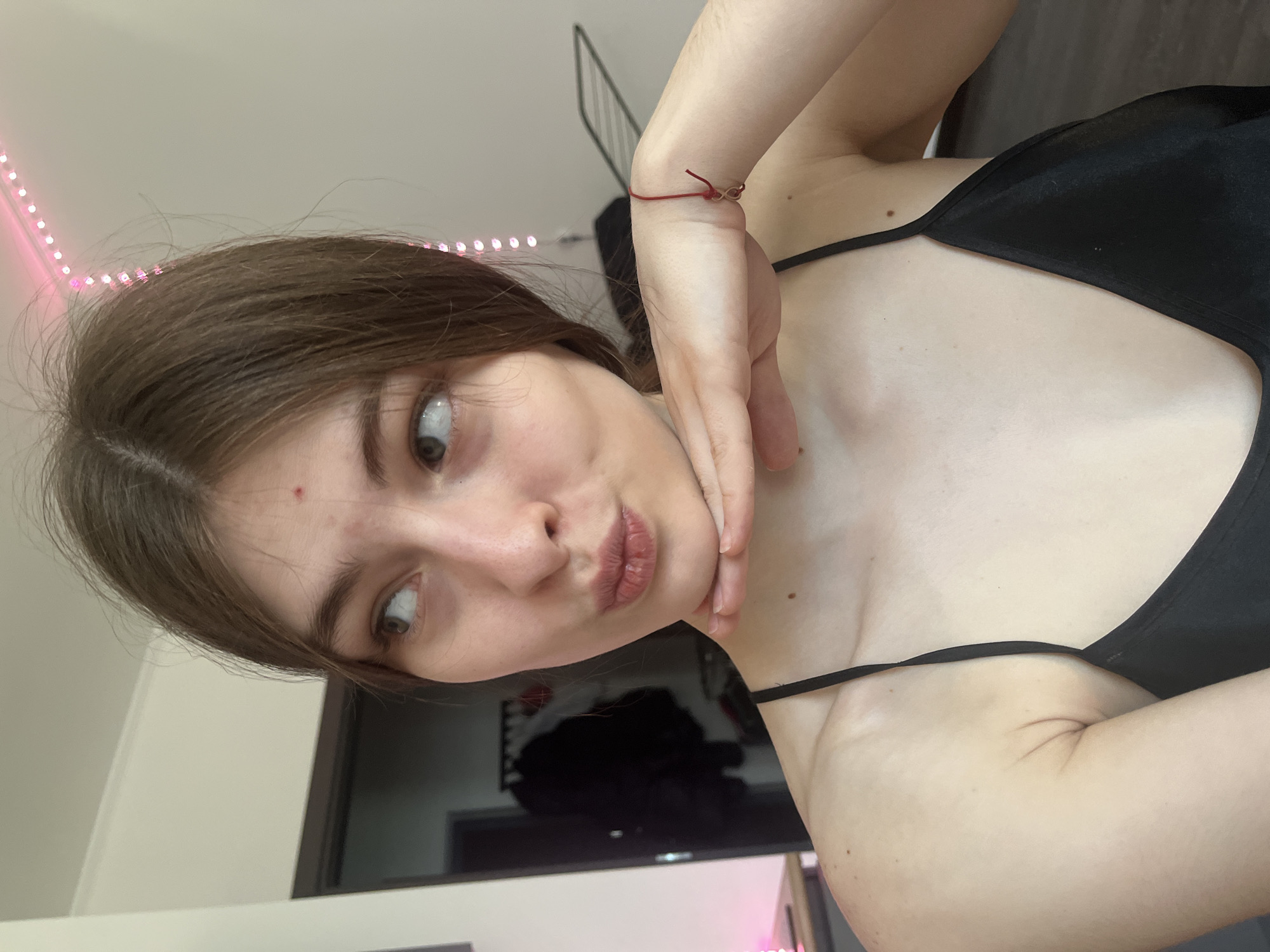 A young person with light skin and brown hair is posing for a selfie. The individual is wearing a black spaghetti strap top. The person's left hand is touching their face. A red bracelet is on their left wrist. A pink light is visible in the upper left corner of the frame. A television or monitor is visible in the background. There is no information to suggest that this image was taken in Saint Petersburg, Russia.