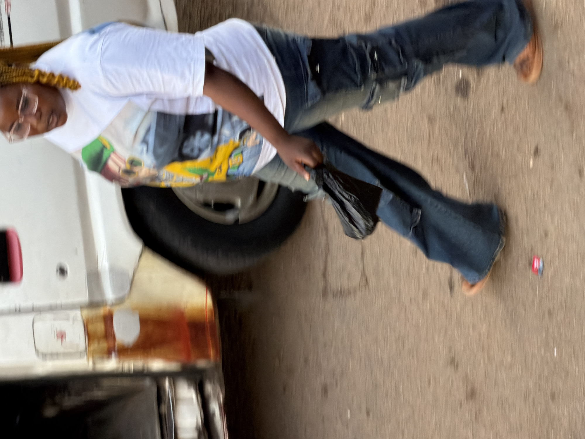 A person walks on a dirt road next to a white van in Bukuru, Nigeria. The person, wearing a white t-shirt with a graphic print and blue jeans, is carrying a black plastic bag. The van appears to be older and shows signs of rust and wear. The ground is a mix of dirt and gravel, with some scattered debris. The lighting suggests it is daytime.