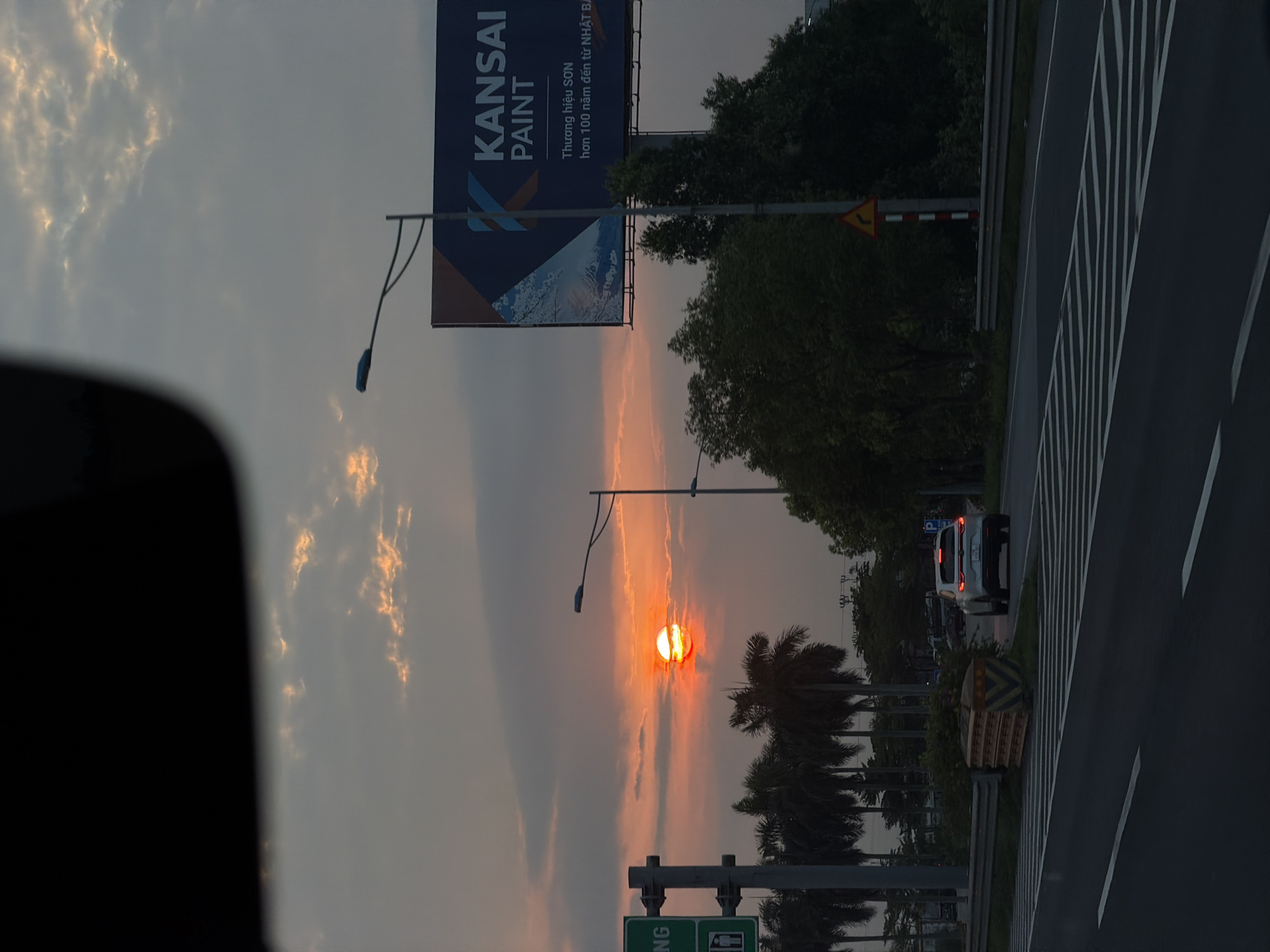 The image shows a sunset viewed from a vehicle in Hải Phòng, Vietnam. A Kansai Paint billboard is visible on the left. The sun is setting near palm trees. A road with lane markings is on the right, with a car driving on it. There is a street light. A road sign is on the bottom left.