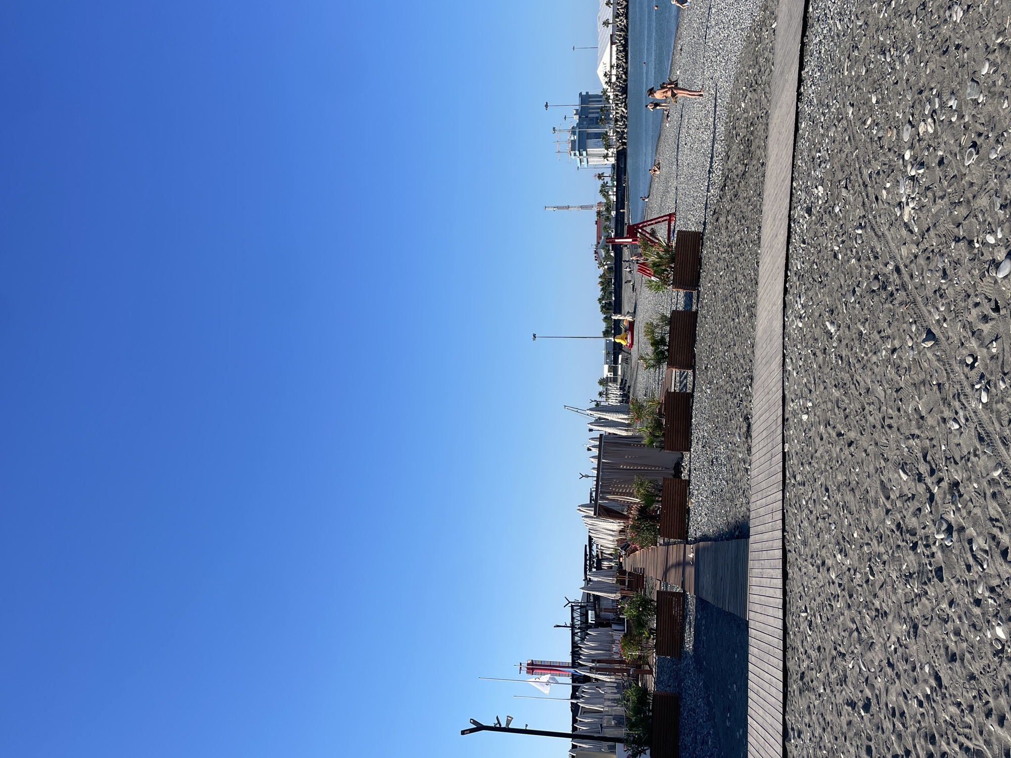 The image depicts a sunny day on a pebbly beach in Sochi, Russia, near a port. The foreground is dominated by a wooden walkway made of planks, leading from a sandy and pebbly beach to a section of the beach lined with rows of closed white beach umbrellas and partitions. Planters with green plants are positioned along the walkway.
Further back, the beach is occupied by a few sunbathers and people walking. In the background, a port infrastructure is visible with cranes and a large building complex. The sea is calm and blue, meeting the clear blue sky. The scene appears to be a developed tourist area within the city.