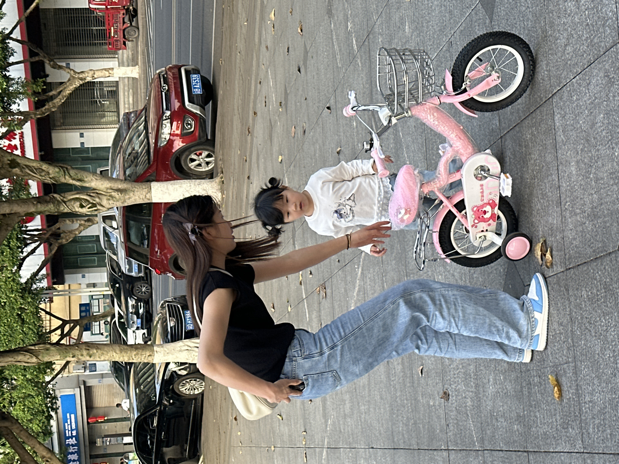 This image, set on a bright day in Jiangjin, China, captures an outdoor scene on a tiled sidewalk adjacent to a street with parked cars. The central subjects are a woman and a young girl interacting near a child's bicycle.
The woman, with long dark hair, wears a black short-sleeved top, light blue wide-leg jeans, and light blue and white sneakers. She carries a light-colored shoulder bag. She is leaning slightly forward with her right arm extended towards the child, appearing to assist or instruct.
The young girl, likely a toddler or preschooler, has dark hair styled in two small pigtails. She wears a white long-sleeved top with a graphic print. She stands beside a vibrant pink child's bicycle, looking towards the woman. The bicycle features a front basket, training wheels, and a pink padded seat, with a red cartoon bear decal on the chain guard.
The background shows an urban street lined with trees and several parked cars. A red SUV with the license plate "渝 B A2539" (Chongqing) is visible behind the woman. Further down the street, a black sedan displays the license plate "沪 DH9R6" (Shanghai). Buildings with commercial ground floors are present, one featuring a blue sign with Chinese characters including "江津" (Jiangjin) and "桂林米粉" (Guilin Rice Noodles), confirming the location context. Scattered dry leaves are visible on the sidewalk. The scene depicts a quiet moment, possibly a parent teaching a child to ride a bike.