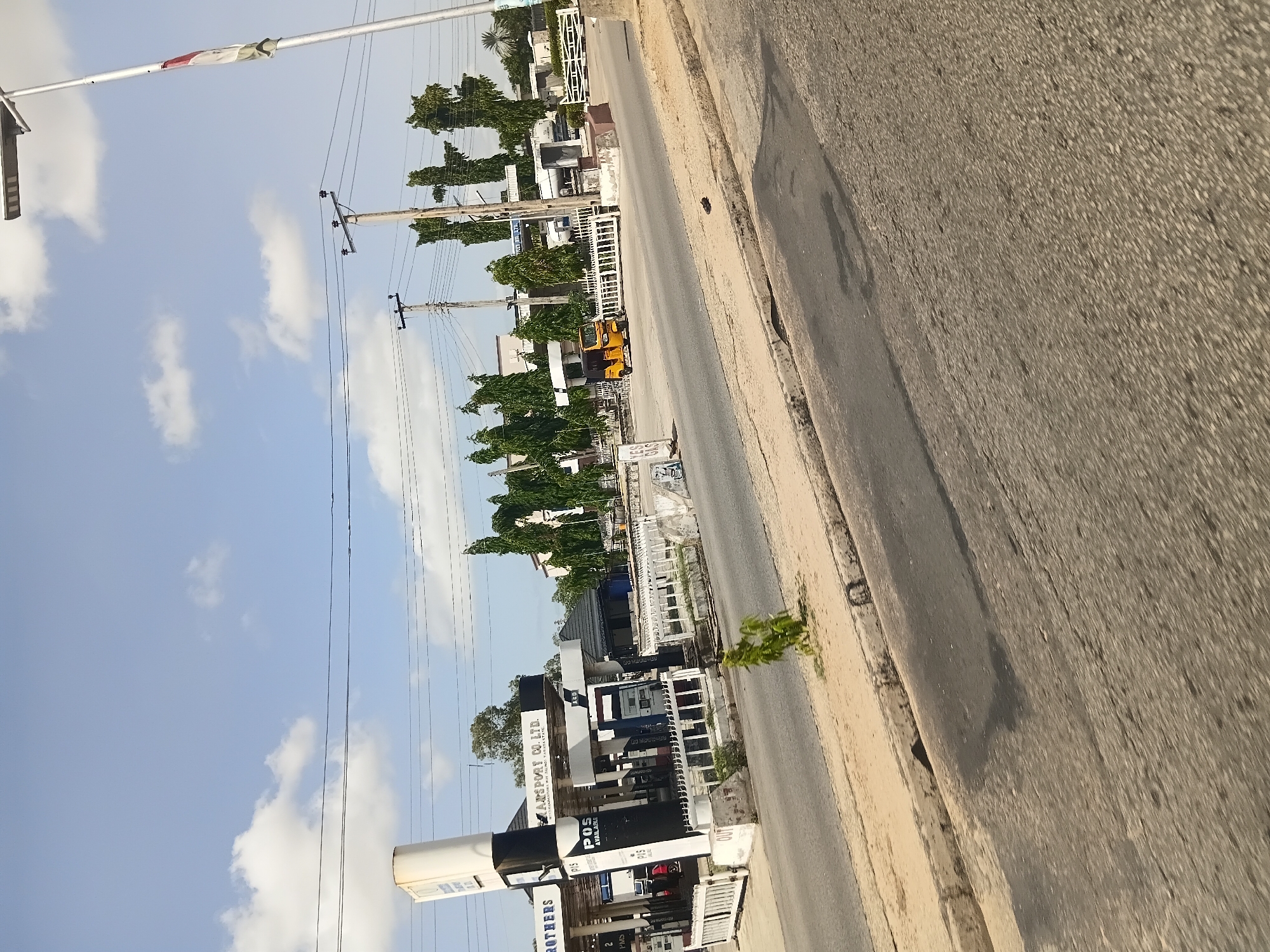 The image captures a street scene in Jalingo, Nigeria, dominated by a gas station and its surroundings under a partly cloudy sky. On the left, a tall, white and black gas station sign is prominently displayed. The gas station itself has a white roof supported by pillars, with multiple pumps visible. Signs indicate "POS AVAILABLE." A yellow three-wheeled auto rickshaw is parked on the asphalt in front of the gas station. A white metal barrier separates the gas station from the road. Power lines run across the sky, supported by utility poles. In the background, trees line the area behind the gas station, and various buildings are visible. A modern street lamp with solar panels stands on the right side of the frame, featuring a flag at the top. The foreground shows a paved road with a sandy shoulder, and a small green plant is growing on the shoulder near the curb. The overall impression is that of a typical roadside commercial area in a Nigerian city.