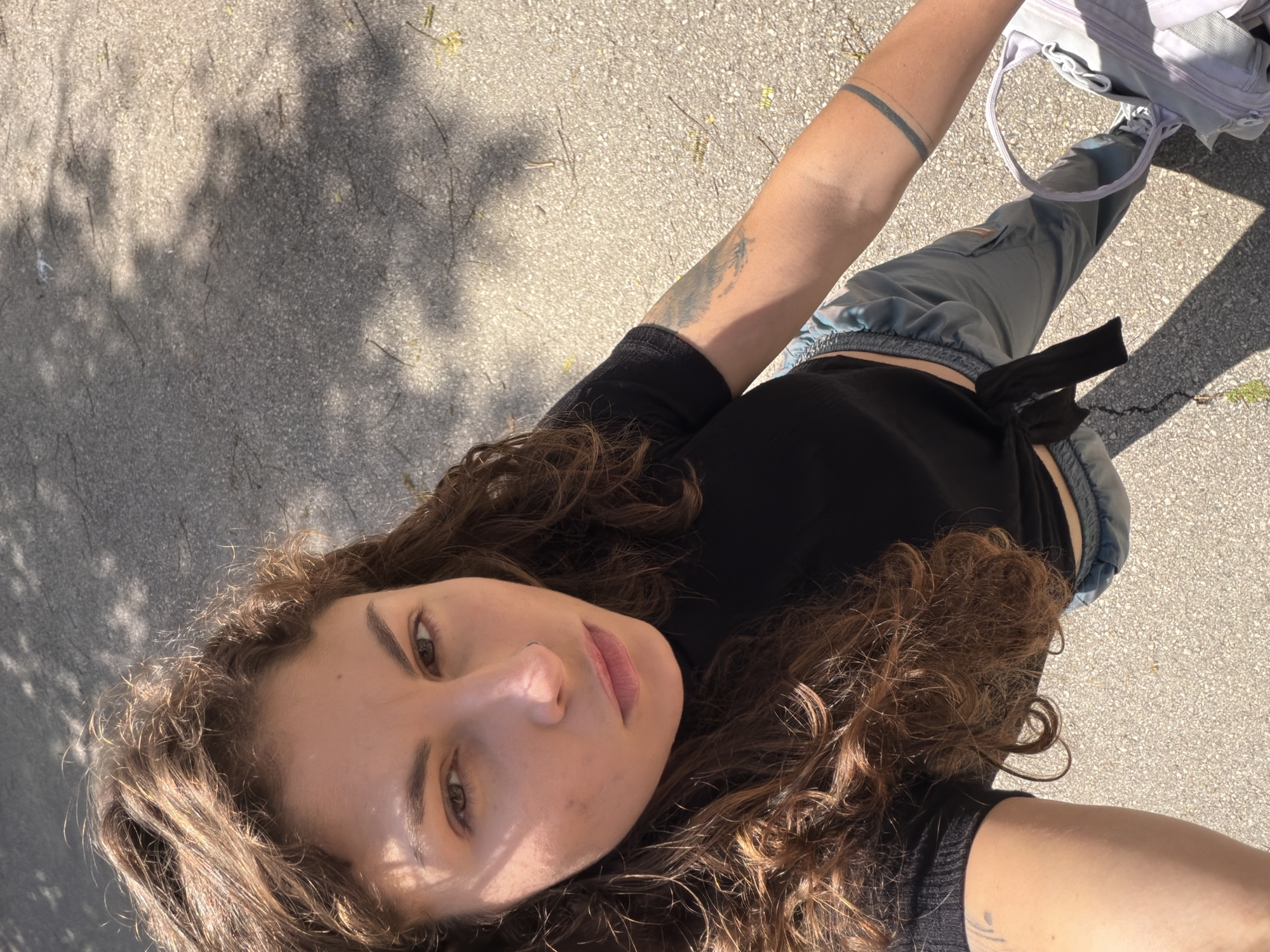 A woman is standing outdoors on a paved surface in Kiryat Bialik, Israel. She is fair-skinned with brown, curly hair. She is wearing a black shirt and light blue cargo pants with a black tie at the waist. A white backpack and white sneakers are visible. The woman has tattoos on her forearms, a double-banded tattoo on her right arm and a tattoo on her left arm. Sunlight filters through trees, creating shadows on the pavement.