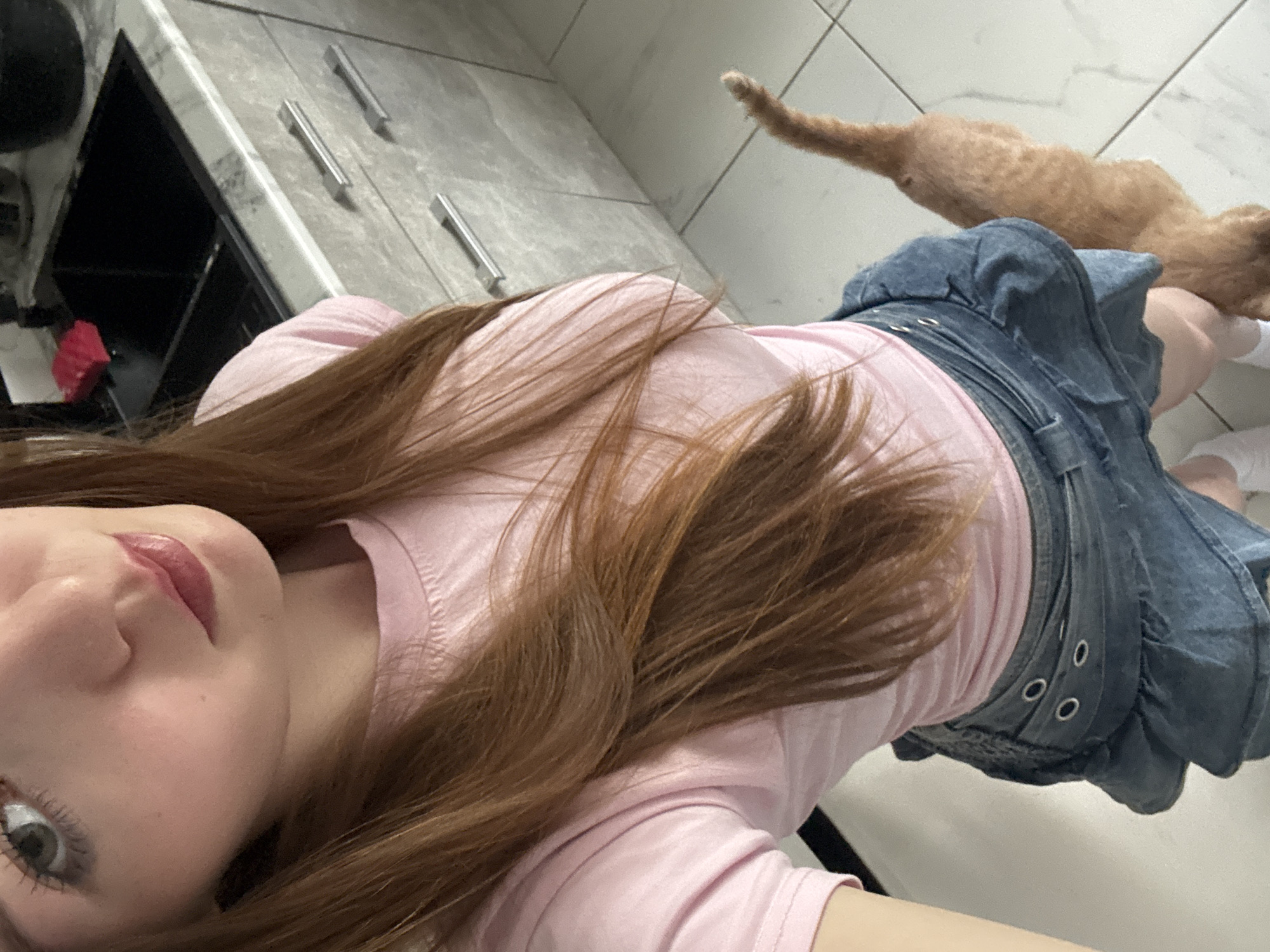 A young woman with long, auburn hair and fair skin, wearing a pink t-shirt and a denim skirt, poses for a selfie in a kitchen. Her lips are glossed, and she has subtle eye makeup. A ginger cat is partially visible in the lower right corner of the frame, its tail extending upwards. The background reveals kitchen cabinets with grey, marble-patterned countertops and stainless steel handles, as well as a black sink. The floor is tiled with a light grey pattern. The lighting suggests an indoor environment, likely in a home. This scene is not identifiable as being specifically in Chaikovsky, Russia, or any other city.