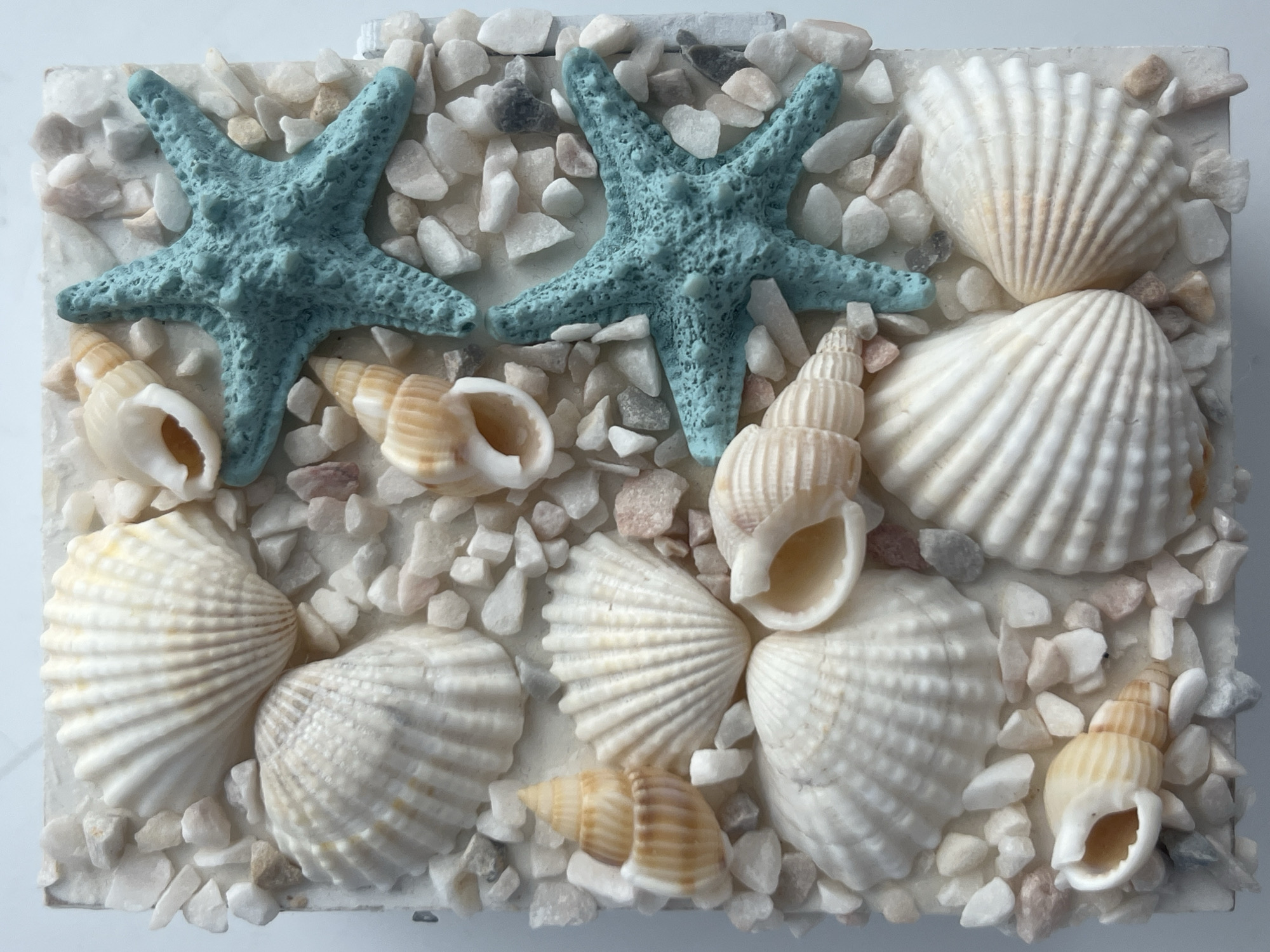 The image displays a close-up, overhead view of a decorative item that resembles a miniature beach scene. The base of the object is white and appears textured, possibly representing sand or a canvas. It is densely covered with small, multi-colored pebbles and various seashells, creating a mosaic-like effect.
Prominently featured are several seashells, predominantly white and cream-colored, with ribbed textures. These include fan-shaped shells and spiral shells. Interspersed among the shells and pebbles are two bright teal-colored starfish. The starfish have a textured surface and are positioned diagonally across the frame. Smaller, tan-colored spiral shells with distinct ridges are also visible, adding variation in color and shape. The arrangement suggests a deliberate artistic composition.
There are no people, animals, or any indication of a specific location within the city of Rybnitsa, Moldova. The scene is static and purely decorative.