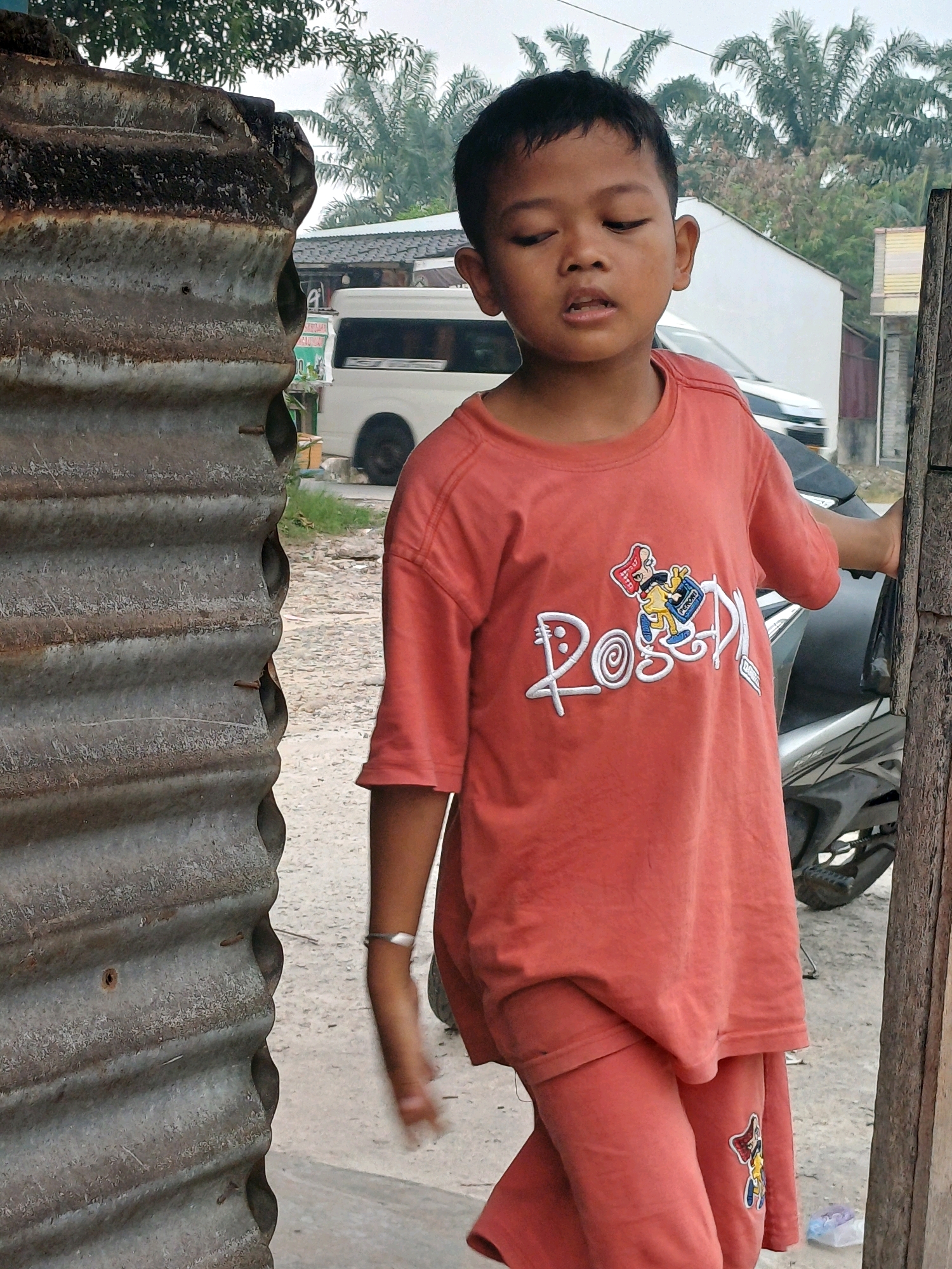 A young boy with dark hair and brown eyes stands outdoors, wearing an orange t-shirt with a cartoon character and white text that appears to read "ROSED". He is also wearing matching orange shorts. A silver bracelet is visible on his left wrist. He is standing next to a corrugated metal wall on the left and a wooden post on the right. In the background, a white van, a motorcycle, and palm trees are visible, suggesting a tropical or subtropical environment. The ground is covered with gravel and dirt. The lighting indicates it is daytime, possibly overcast. The location is identified as the city of Dumai, Indonesia.