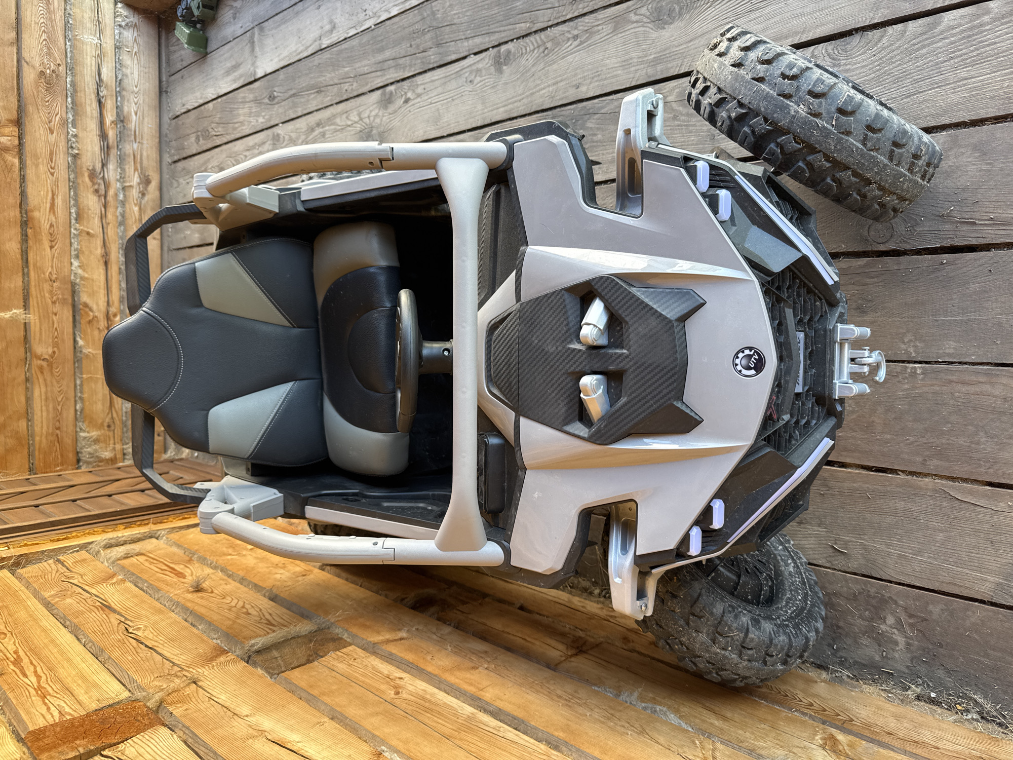 A top-down, slightly angled view captures a grey and black toy UTV parked on a wooden deck next to a log wall. The UTV features two seats with black and grey upholstery, a black steering wheel, and a silver roll cage. The front of the UTV has a grey body with carbon fiber accents and two headlamp-like features. A detached tire rests to the right of the vehicle on the wooden planks. The log wall to the left is composed of rough-hewn, natural logs. The wooden deck is made of weathered planks. The scene is outdoors, suggesting a rural or natural setting. There are no people or animals visible. The UTV appears to be stationary.