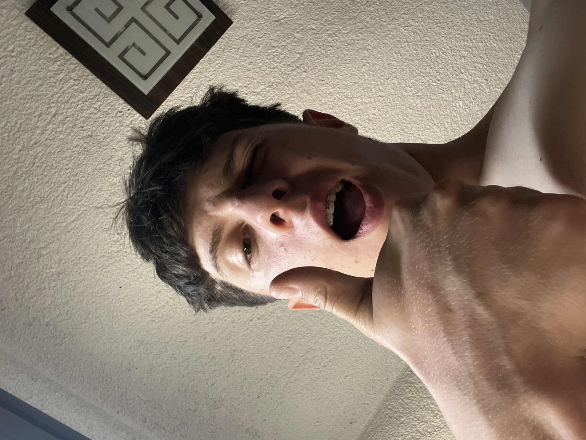 A low-angle indoor shot captures a young person from the chest up, positioned towards the top of the frame. Their head is angled down towards the camera, slightly tilted to the right. They have dark, slightly disheveled hair. Their right eye is visible and open, while the left eye is squinted or closed. Their mouth is wide open, revealing braces on their upper teeth. The person's right hand is raised, with fingers partially obscuring the lower right side of their face, near their jawline. The skin tone appears fair, with some blemishes visible on the cheek and forehead.
The background consists of a textured, off-white ceiling. In the top left corner of the frame, part of a square light fixture is visible, featuring a dark brown frame with a light-colored, labyrinthine pattern. The lighting casts shadows, particularly on the lower part of the person's face and neck, and highlights their forehead and cheekbones. No text is visible in the image. The time of day cannot be determined, but the indoor setting suggests artificial lighting or light filtering through from outside. The location context provided is Zlatograd, Bulgaria.