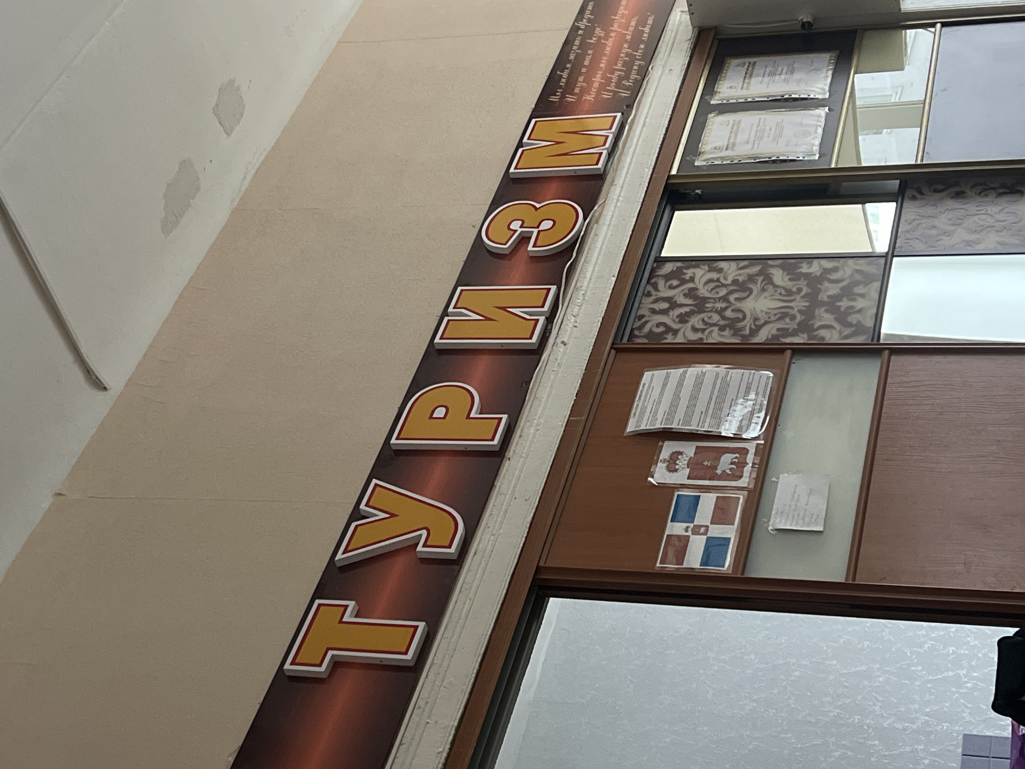 This is a slightly angled, close-up shot focusing on a storefront or office in what appears to be Chaikovsky, Russia. A sign vertically positioned on the facade prominently displays the word "ТУРИЗМ" in large, blocky, red and yellow outlined Cyrillic letters. The sign is affixed to a wall painted a light beige color, which shows some imperfections.
Adjacent to the sign is a storefront window or entryway area. Within this space, certificates are displayed, along with sheets of paper, and two small decals resembling regional flags or symbols. One such decal features a white bear and a crown, while the other is a blue and white flag. The window provides a glimpse into the interior, revealing a textured white surface, possibly another wall or a patterned curtain.
The image captures a mundane urban scene in Chaikovsky, Russia, with a focus on the advertising of a tourism business.