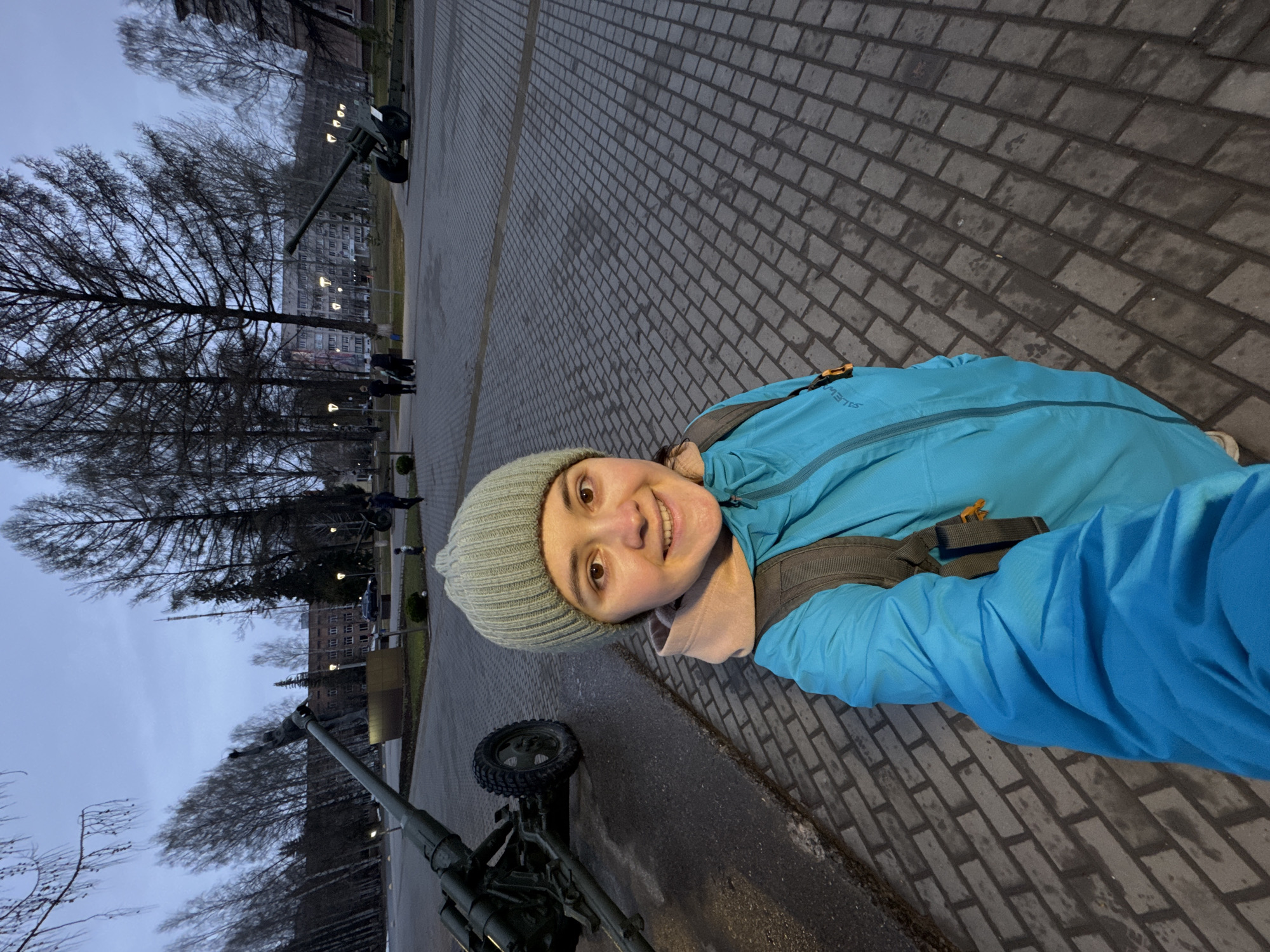 A woman in a blue jacket and a grey beanie smiles at the camera, likely taking a selfie. She stands on a brick pavement. In the background are bare trees and an artillery gun. Based on the appearance of the artillery and the surrounding architecture, this photograph likely depicts a scene at the Memorial Complex of Military Glory in Izhevsk, Russia.