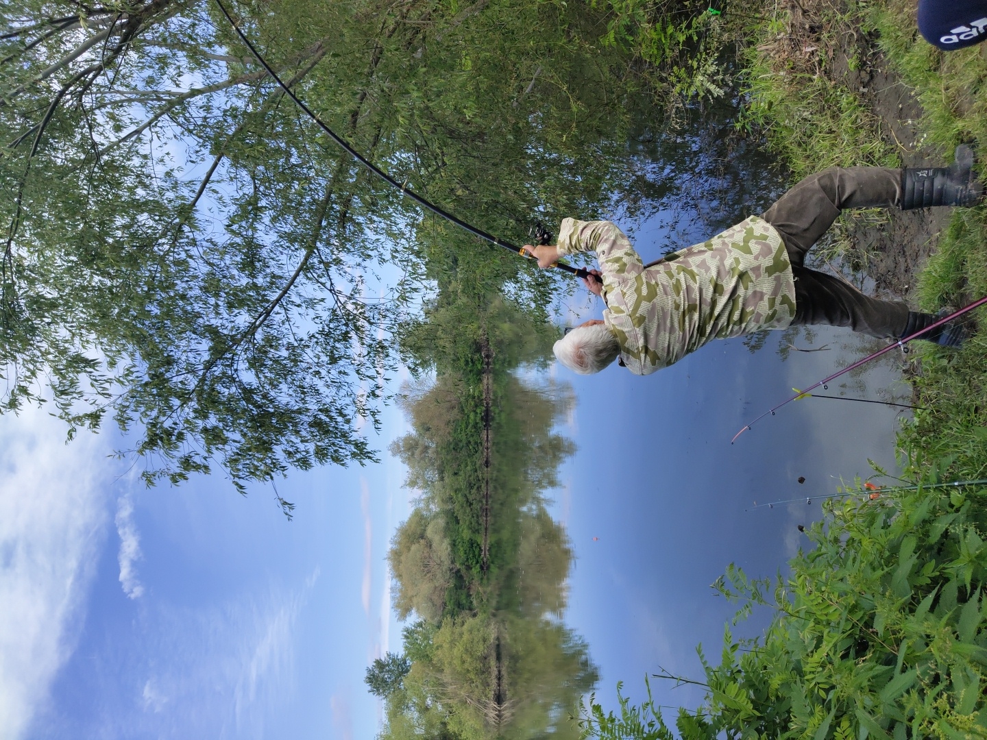 An elderly man with graying hair is fishing on the bank of a calm body of water, possibly a river or lake, with lush green vegetation on the opposite bank and a tree overhanging the water on the right. The sky is blue with scattered white clouds. The man is wearing a camouflage patterned shirt, brown trousers, and black boots. He is holding two fishing rods, one of which is bent significantly, indicating he is actively engaged in fishing. The water reflects the sky and surrounding trees. Several other fishing rods are planted in the ground near the man, suggesting he is in a dedicated fishing spot. The location is identified as Krushare, Bulgaria.