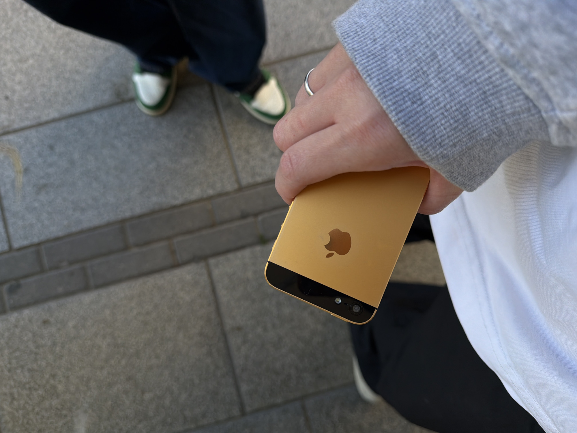 A close-up, high-angle outdoor shot captures a person's hand holding a gold-colored iPhone 5S, with the iconic Apple logo visible on its back. The phone is positioned with its camera module facing upwards and its screen angled away from the viewer. A simple silver band ring adorns one of the fingers holding the device. The person is dressed in casual attire, featuring a grey ribbed sleeve, likely from a sweatshirt or hoodie, and a white t-shirt.
In the blurred background, the lower legs and feet of another individual are visible. This person wears dark-colored pants and white sneakers with distinct green trim. Both individuals appear to be on a paved outdoor surface, consisting of large, rectangular grey paving stones, characteristic of a sidewalk or urban pedestrian area.
The scene is brightly lit, indicating it is daytime with clear weather. No strong shadows are cast. The location context provided indicates the image was captured in Tel-Aviv, Israel. No faces are visible, so emotions cannot be discerned.