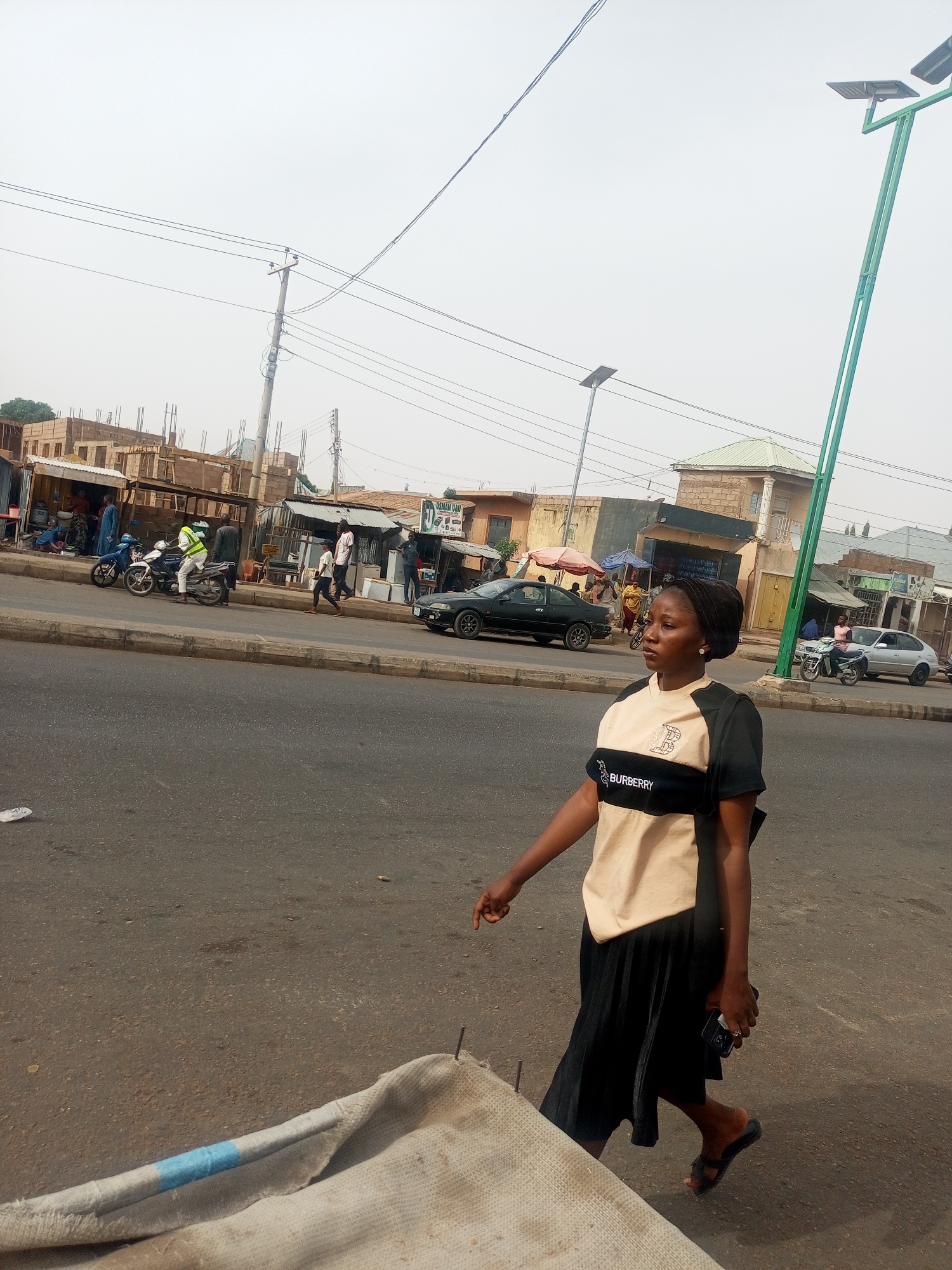 The image presents a daytime street scene in Tempure, Nigeria, under an overcast or hazy sky. In the foreground, a woman with dark skin walks along the edge of a paved road, her gaze directed towards the right. She is dressed in a short-sleeved, two-toned shirt, featuring beige across the chest and black on the shoulders and lower portion, with "BURBERRY" and a "B" logo clearly visible. She pairs this with a black pleated skirt and black sandals, carrying a mobile phone in her left hand.
The background reveals a bustling urban environment. Numerous people are visible on the sidewalks and near roadside structures, some walking and others gathered. A person wearing a high-visibility vest is seated on a motorcycle. Several cars and additional motorcycles are present on the road and parked along the curb.
Along the street, various buildings and makeshift structures line the road. Some buildings are visibly under construction, indicated by exposed rebar. There are also smaller shops or market stalls with tarps and metal roofs. Utility poles with multiple power lines dominate the upper part of the scene, alongside modern streetlights, some equipped with solar panels. A sign in the distance includes the text "JUSHAN DAU". A large piece of light-colored fabric or tarp lies partially in the bottom left foreground. The scene captures a typical day in a vibrant, developing urban area.