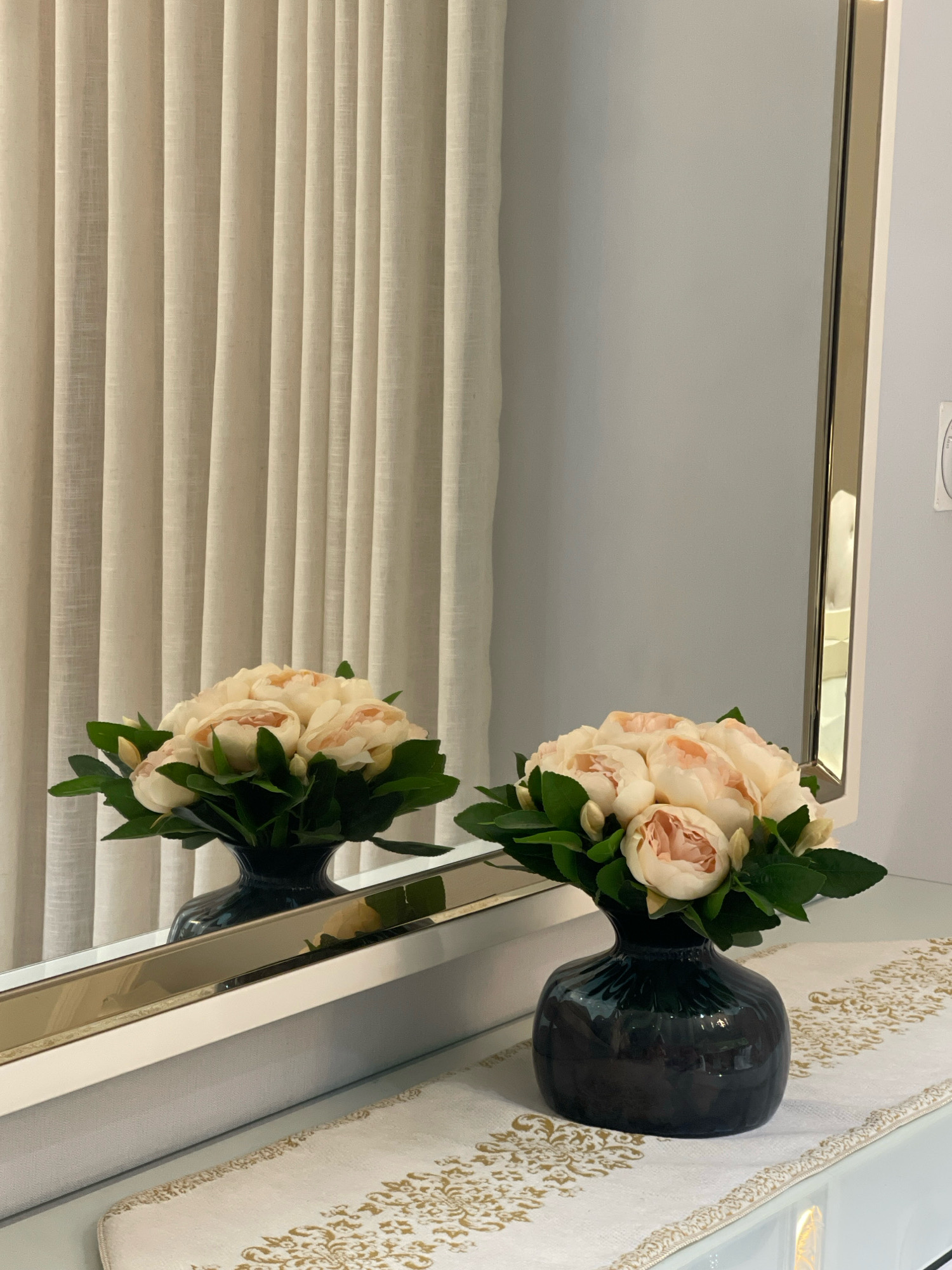 A bouquet of light pink roses with green leaves is arranged in a dark, glossy vase. The vase is placed on a white table runner with an ornate gold pattern. The scene is reflected in a mirror, which also shows beige vertical curtains in the background. The overall setting suggests a domestic interior, possibly in a room in Jabriya, Kuwait. The image is well-lit and focuses on the floral arrangement and its reflection. There are no people or any discernible actions taking place.