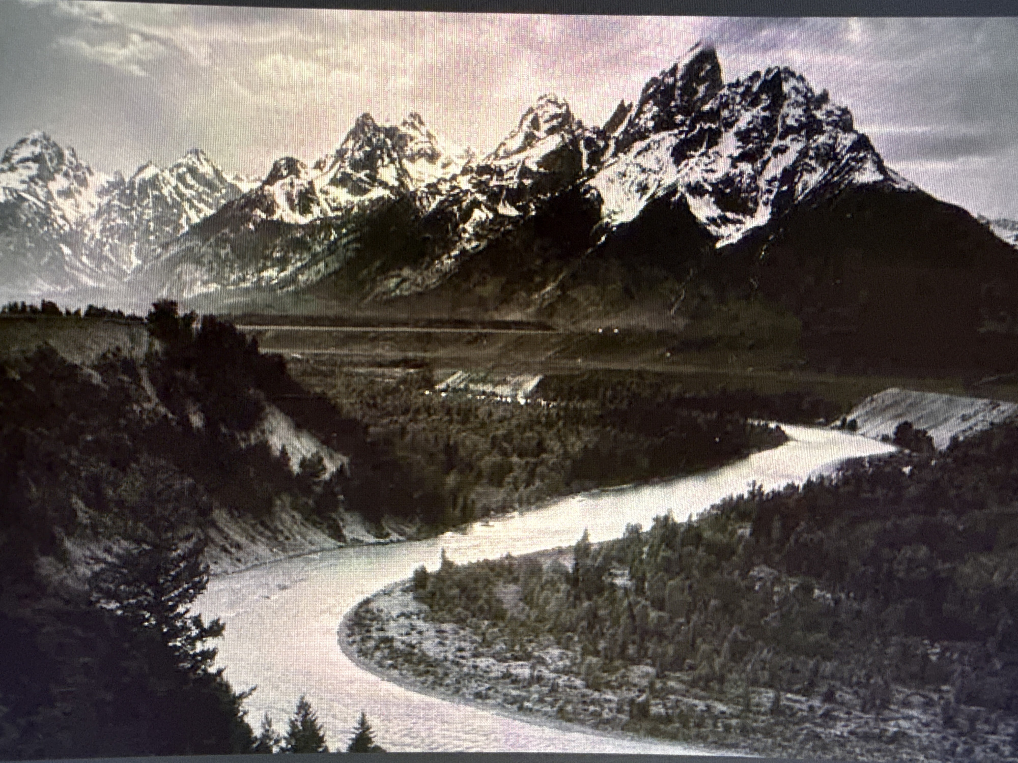 A monochrome photograph shows a river curving through a valley towards a range of snow-capped mountains. Dense trees cover the valley floor. The scene appears to be in a rural, mountainous region. There is no indication of Sofia, Bulgaria in the image.