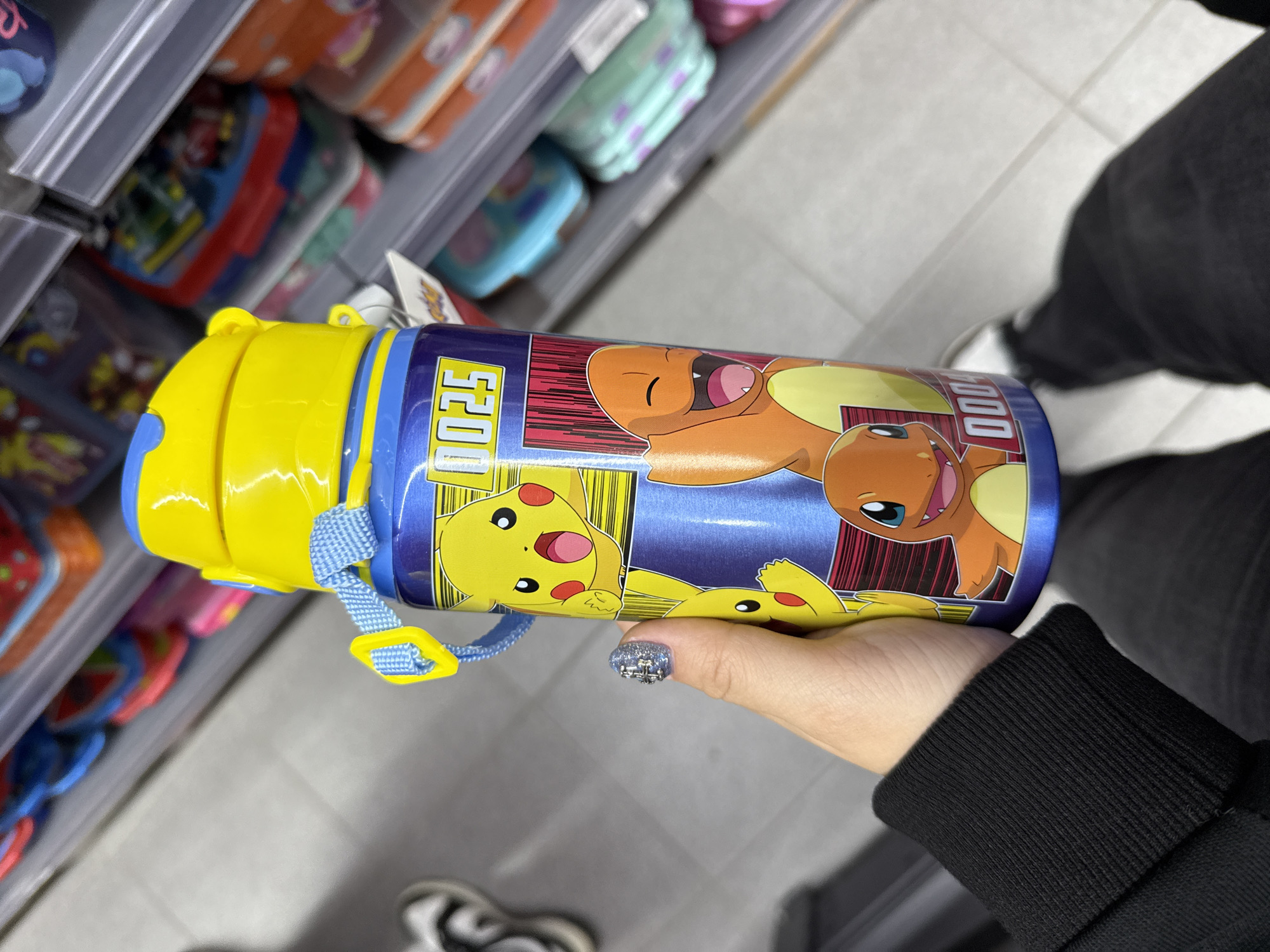 A hand holds a blue and yellow water bottle with Pokémon characters Pikachu and Charmander printed on it. The bottle has a yellow lid and a blue strap. The hand has a fingernail painted with a sparkly design. The person holding the bottle is wearing a black long-sleeved shirt and a white and black shoe is visible in the lower left corner. Shelves filled with various colorful items are visible in the background. The photo appears to be taken indoors. There is no evidence of it being taken in Madrid, Spain.