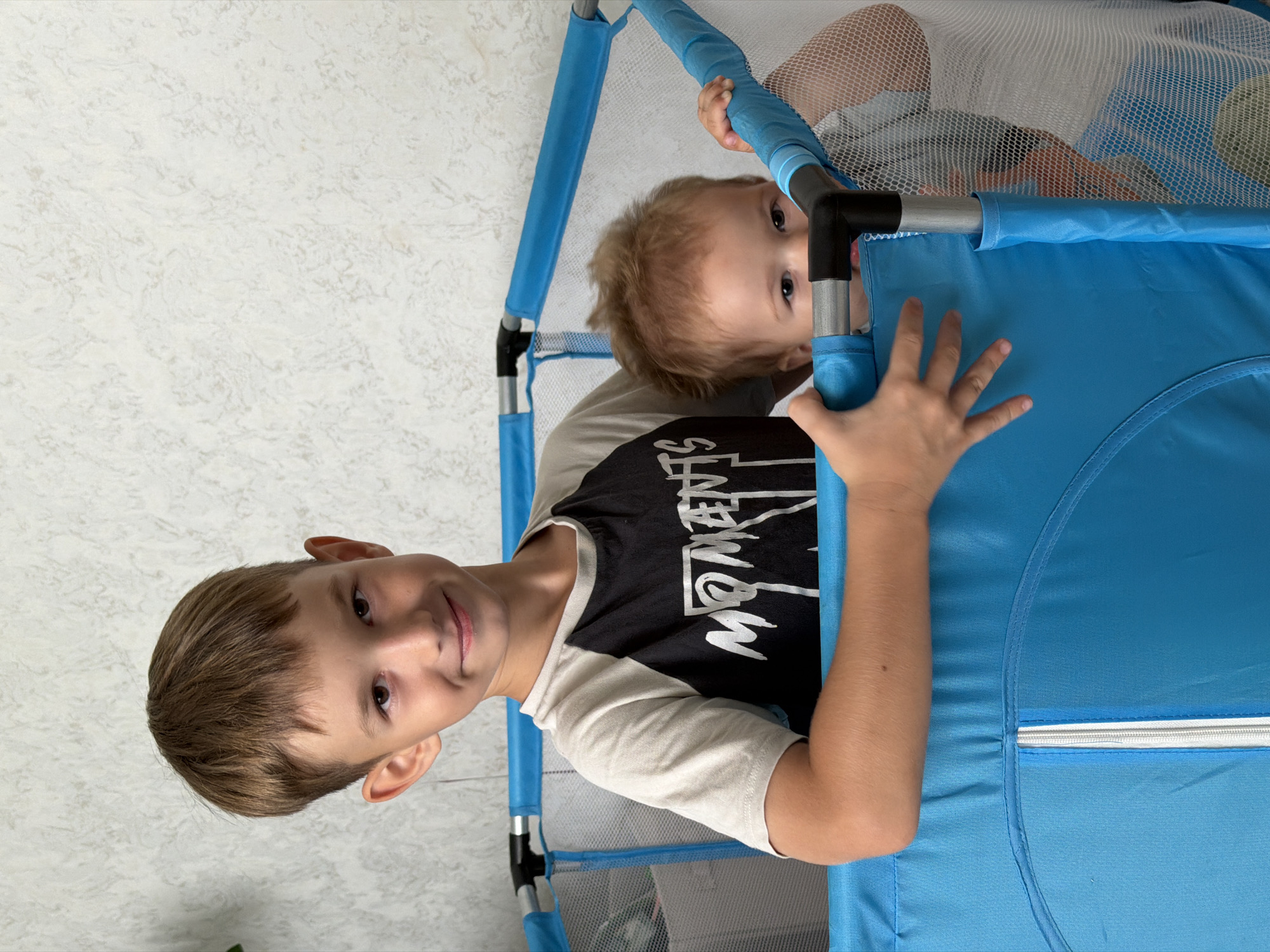 Two young boys are in a blue playpen in an indoor setting. The older boy, on the left, is wearing a t-shirt with a black design and is smiling at the camera. The younger boy, on the right, is peeking over the edge of the playpen and looking at the camera. The background is a textured, light-colored wall. The image does not contain information that would allow for its location to be identified as Kazan, Russia.