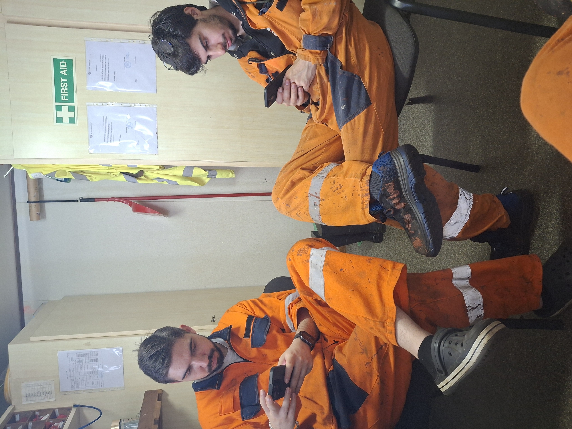 Two individuals wearing orange work jumpsuits are seated indoors. The man in the foreground is wearing black Crocs and looking at his phone. The second man in the background is wearing dark sneakers and is also looking at his phone. Behind them is a wall with a "First Aid" sign, documents, and a yellow vest hanging. The specific location in Cuxhaven, Germany cannot be determined from the image.