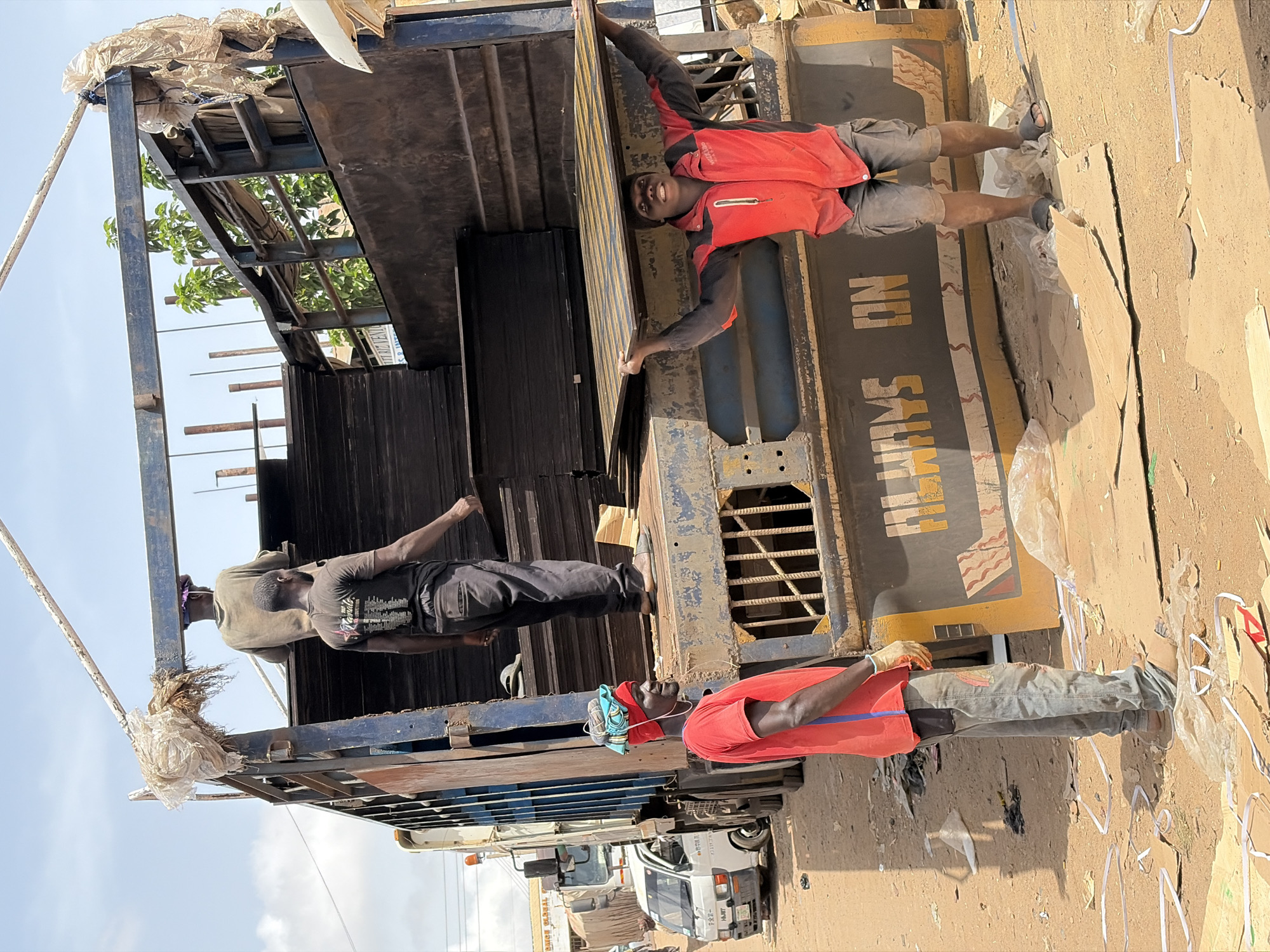 In Gura Top, Nigeria, three men are unloading black boards from a truck. One man is in the truck bed, reaching for the boards, another stands on the truck's side, and a third man is on the ground. The truck is parked on a dirt road in what appears to be a residential or commercial area, evidenced by buildings and other vehicles in the background.