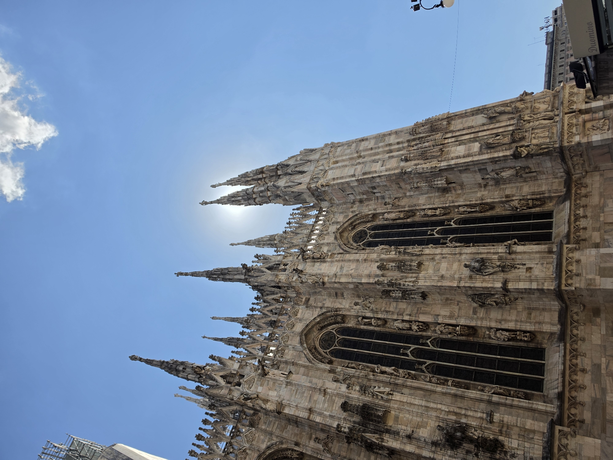 The image shows a low-angle, wide shot of the Duomo di Milano (Milan Cathedral) in Milan, Italy, on a clear, sunny day. The sun is positioned behind the upper spires of the cathedral, creating a bright halo effect and causing some lens flare. The architectural details of the cathedral's facade, including its numerous spires, Gothic windows, and intricate sculptures, are prominently visible. The sky is a vibrant blue with a single, small white cloud near the top right. In the foreground, the lower section of the cathedral is made of light-colored stone. To the far right, a lamppost with a white lamp is partially visible, and a building with signage that reads "Duomino" can be seen. The overall scene captures the grandeur and intricate stonework of this famous Milanese landmark.