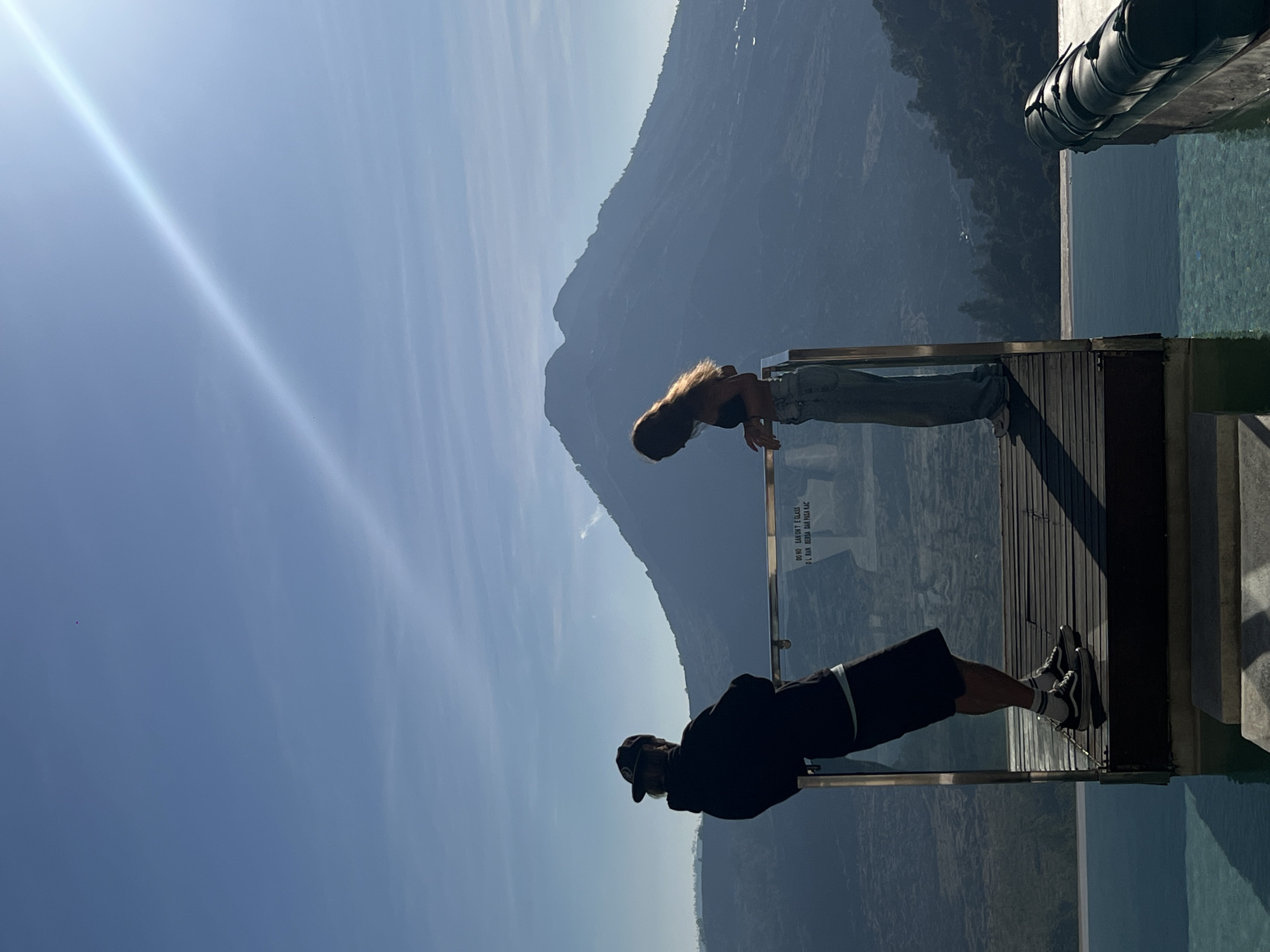 The image displays two individuals on a wooden viewing platform with a glass railing, overlooking a vast mountainous landscape and an infinity pool in Kintamani, Indonesia. The person on the left, viewed from behind, wears a dark baseball cap, a dark hoodie over a light t-shirt, dark shorts, white socks, and black Vans-style sneakers. They lean with forearms on the top rail of the glass barrier. The person on the right, seen in profile, has long, light-brown hair and wears a dark bra-top with light blue wide-leg jeans and white sneakers, also leaning on the glass railing. Text is visible on the glass barrier, including "DO NO EAN ORT E GLASS" and "D L RAN BERPA DAR PASA KAC."
The foreground features a turquoise infinity pool that seamlessly merges with the distant horizon. The background is dominated by a large, dark blue mountain, consistent with Mount Batur, under a clear, bright blue sky with minimal cloud cover. A strong, diagonal lens flare extends from the upper right, indicating direct sunlight. A wide valley, characterized by dark green and blue tones of vegetation and distant land features, lies between the platform and the mountain. A dark, cylindrical object secured with straps is partially submerged in the pool on the far right.