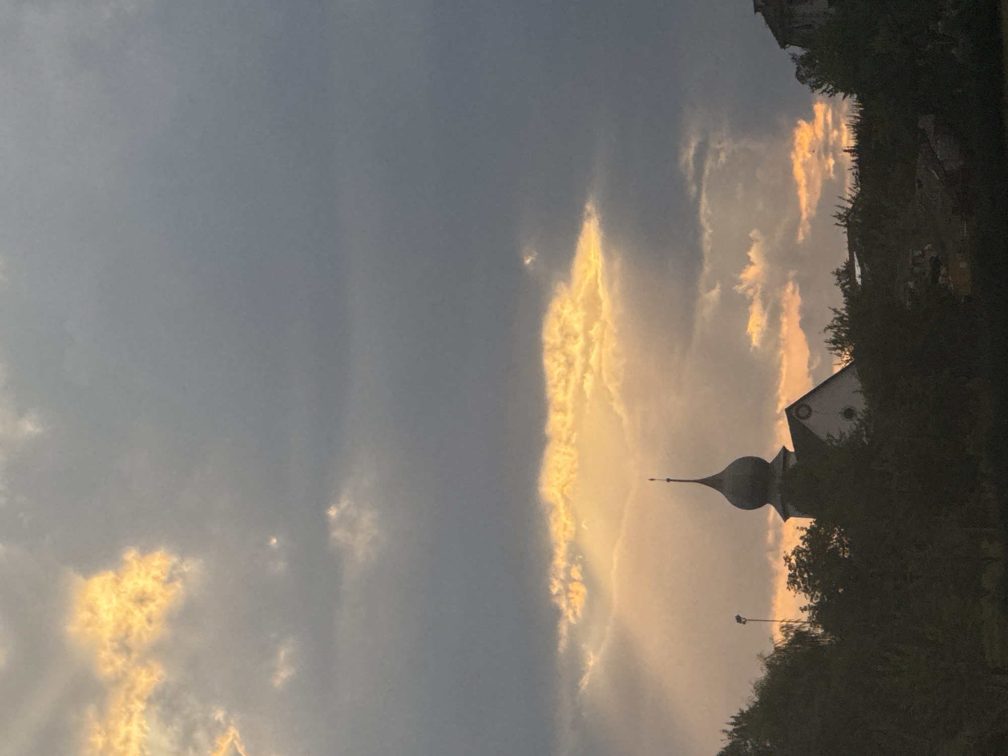 The image captures a dramatic sunset over the town of Petit-Val, Switzerland, featuring a church with a distinctive dome. The sky is filled with a mix of dark, ominous clouds and patches of vibrant, golden light breaking through, creating a striking contrast. The sun's rays are visible, illuminating the clouds from behind. In the foreground, silhouetted trees and the rooflines of buildings create a dark band across the lower portion of the frame, obscuring much of the ground level. The church's spire is prominent, rising above the trees and catching the last rays of the setting sun. To the left of the church, a tall pole with a light fixture is also visible. The overall scene is one of natural beauty and a sense of impending twilight.