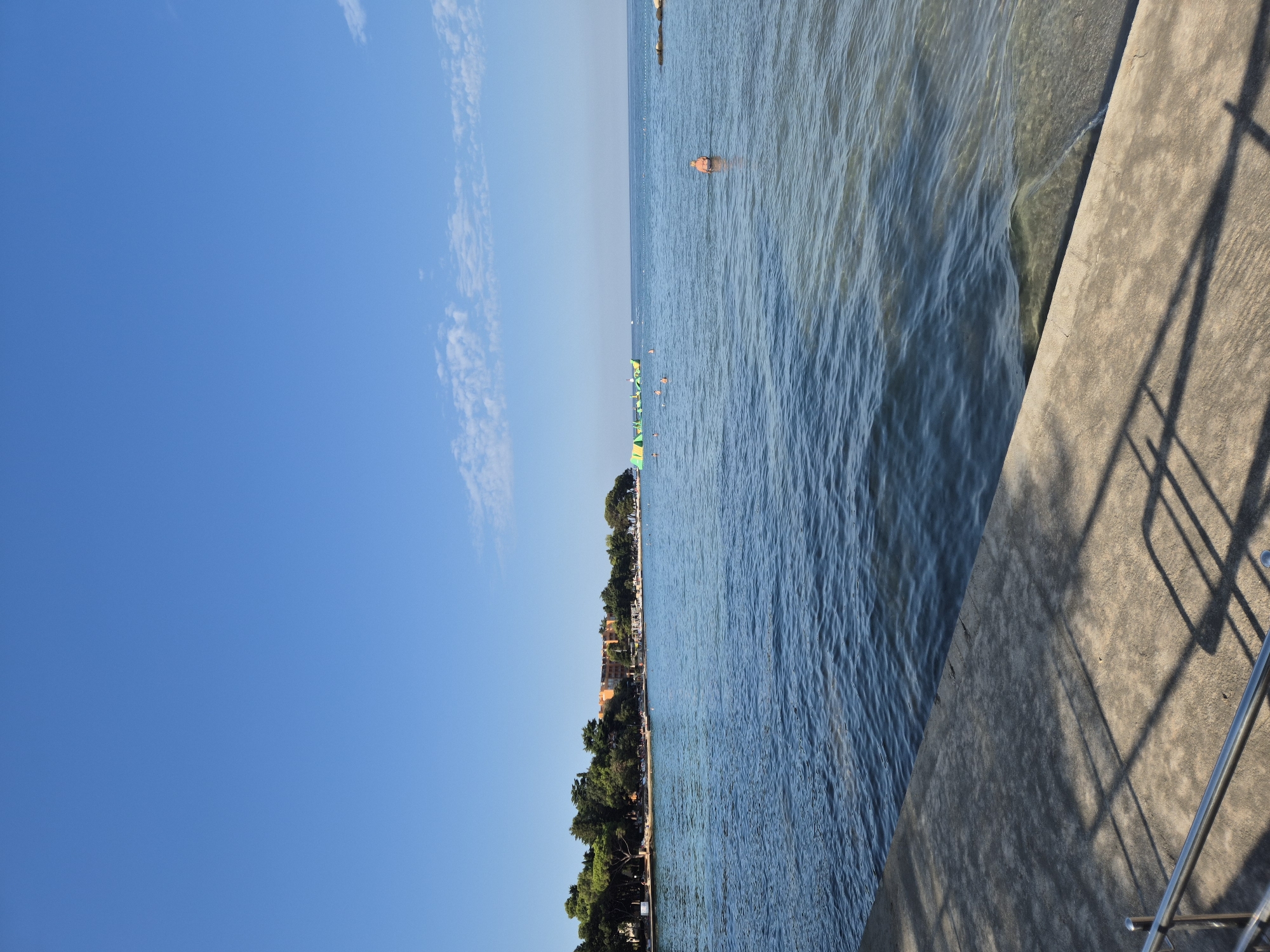 The image shows a serene coastal scene under a clear blue sky. In the foreground, a concrete embankment with metal railings casts long shadows from an unseen structure. The embankment slopes gently down to the clear, blue water of the Adriatic Sea. Small waves lap at the shore, and the seabed is visible in the shallow areas.
In the middle ground, the sea stretches towards a treelined coastline. A few people are visible swimming or wading in the water, and a large inflatable water park structure, colored green and yellow, is anchored offshore. A single person in an orange life jacket is swimming further out. Along the coastline, a strip of sandy beach is lined with trees and a few buildings, including a multi-story orange hotel.
The background is dominated by the vast expanse of the blue sky, with a few wispy white clouds scattered near the horizon. The overall impression is one of a peaceful summer day at a beach resort. The location is identified as being in Monterol, Croatia.