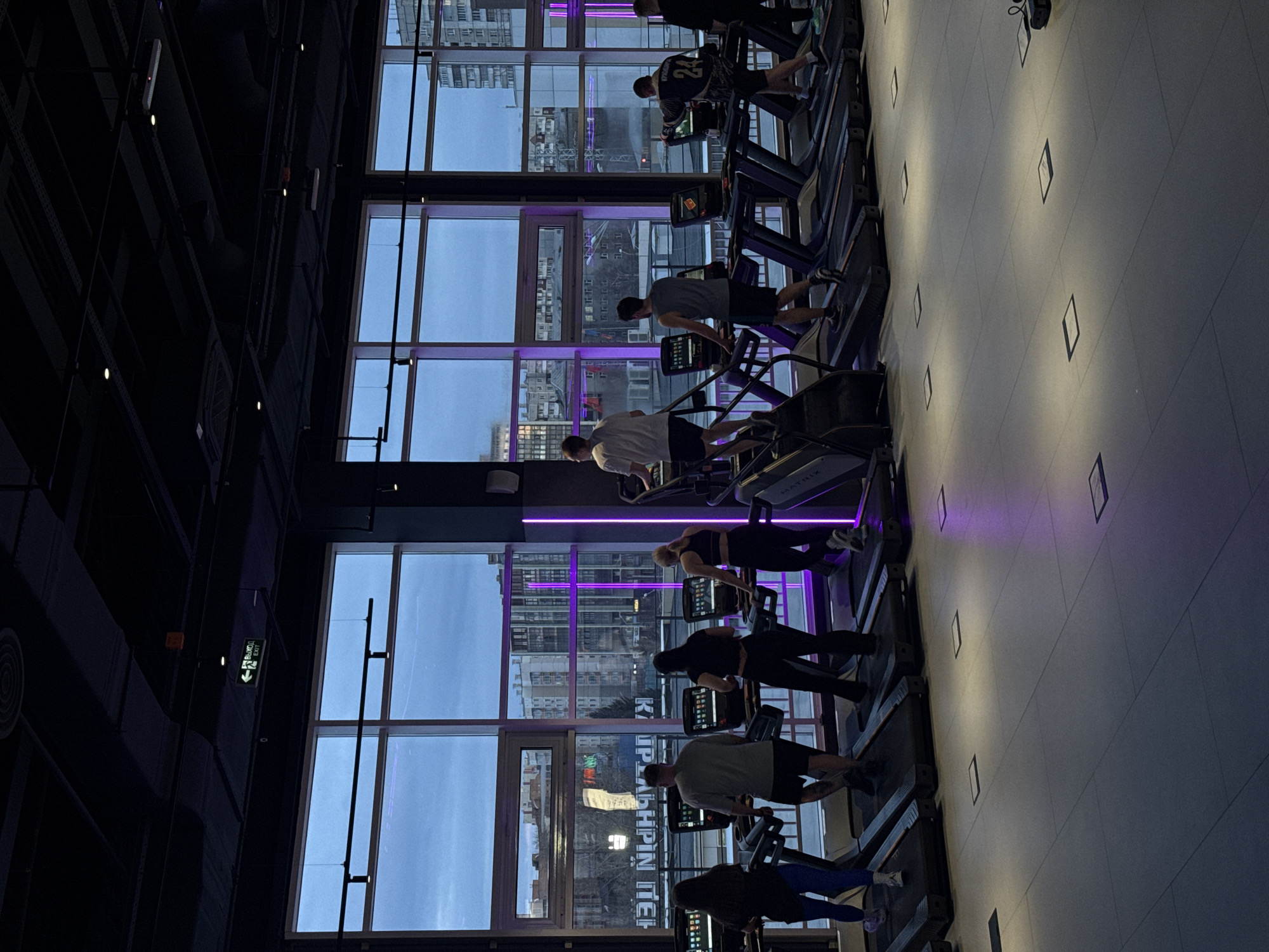 This image captures the interior of a modern gym's cardio section in Krasnoyarsk, Russia, during what appears to be the early evening or dusk. A row of individuals, primarily adults, are actively exercising on treadmills or stair climbers, facing a large expanse of floor-to-ceiling windows.
The scene is illuminated by a mix of indoor lighting and the ambient light from outside. Distinctive purple neon strip lights run along the window frames, casting a vibrant glow that contrasts with the cooler, dusky blue sky visible through the windows. The ceiling features exposed industrial elements and various light fixtures. The light-colored floor is dotted with recessed square lights, contributing to the contemporary atmosphere.
Beyond the windows, an urban landscape of buildings is visible, subtly reflected in the glass. Notably, the reflections include Cyrillic text, with "ТЦ ЛАБИРИНТ" (TC Labyrinth) and "KРАСНОЯРСК" (Krasnoyarsk) clearly discernible, confirming the location.
Among the subjects, one person wears a jersey with the number "24" on their back. The brand "MATRIX" is visible on the side of some of the cardio machines. On a dark wall to the left, a green "ВЫХОД" (EXIT) sign with an arrow points towards an exit. The overall ambiance is focused and energetic, characteristic of a fitness facility.