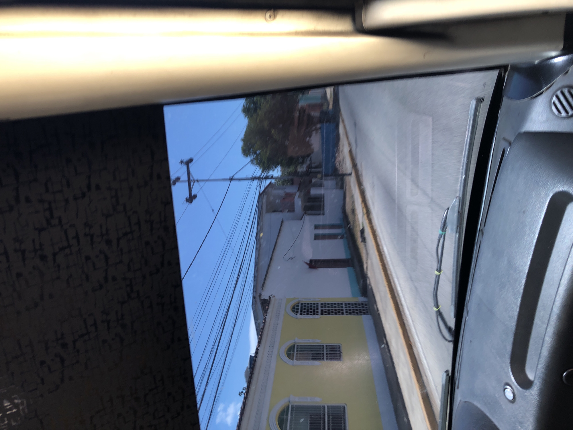 The image, captured from the interior of a vehicle looking outwards, depicts a sunny street scene. On the left side of the frame, interior elements of the vehicle are visible: a bright, illuminated overhead panel at the top, and a dark, textured surface, possibly a wall or fabric, below it. On the right, parts of a dashboard, including an air vent and dark plastic panels, are visible along the window's edge.
Through the vehicle's window, a clear blue sky occupies the upper portion, traversed by numerous utility lines. A tall utility pole stands on the left, supporting these lines. Below, a street features buildings with residential characteristics. A prominent building with a light yellow facade, adorned with arched windows and decorative grilles, is visible in the foreground. Above it, another building with white or light-colored walls and a flat roof extends further into the distance, partially obscured by green trees. A paved road or street runs parallel to the buildings, with a sidewalk or curb visible. A windshield wiper blade is present on the right side of the window, along with bundled wires, confirming the forward-looking perspective from the vehicle.
The scene suggests bright daylight, likely morning or afternoon, with clear weather. No individuals are present. The architectural style and infrastructure align with a typical street environment in a location like Tacarigua, Venezuela. No visible text is discernible within the image.