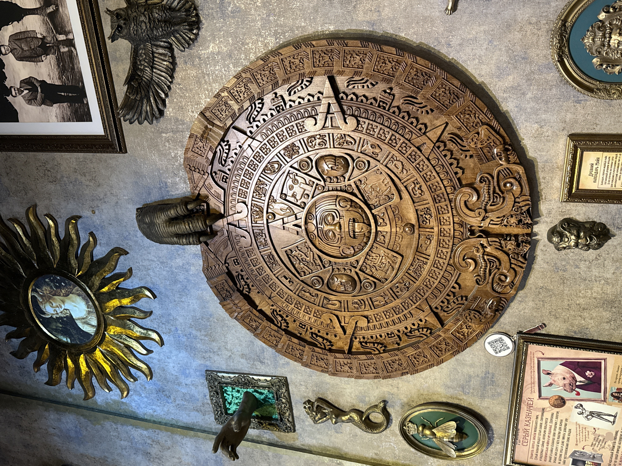 The image shows a wall adorned with an eclectic collection of objects, suggesting a themed decor or exhibit. The centerpiece is a large, intricately carved wooden disk resembling an Aztec or Mayan calendar stone. To the left is a sunburst mirror with a painting inside, a bronze owl sculpture, and a framed black and white photograph. Below the wooden disk are several small framed items, including a green framed object with a bronze hand sculpture, a bronze mermaid figurine, and a framed bee. To the right of the disk are a small framed text, a framed picture of a pig in a suit labeled "GRAY TREASURER", and a decorative circular frame. There is also a QR code near the center of the image. The room has textured, possibly painted, walls. There is no verifiable information about the location being in Moscow, Russia.