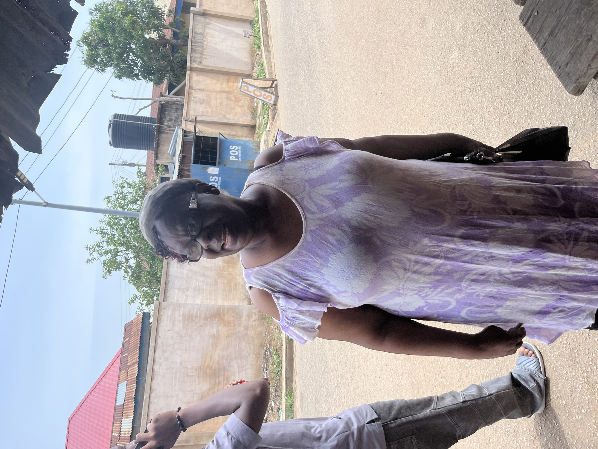 A woman stands on a paved road in Jos, Nigeria. She wears glasses and a light purple and white patterned dress with ruffled sleeves, and she holds a black purse. To her right, a partially visible person wearing a light grey shirt and grey pants stands with one foot visible. The background shows buildings, a water tank, trees, and a "POS" sign. The overall lighting suggests a bright, outdoor setting.