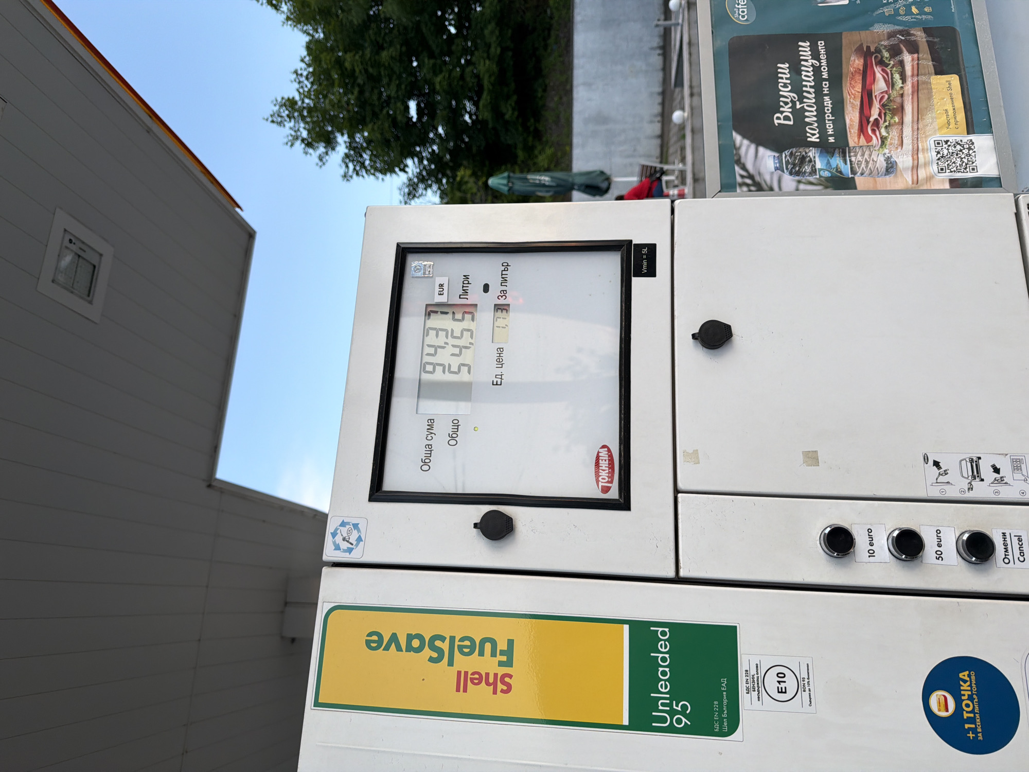 An image, rotated counter-clockwise, depicts a white fuel pump at a Shell gas station under a clear blue sky. The scene is outdoors during the daytime, with green trees visible in the background and part of a white building to the left.
The central subject is the fuel pump's digital display, showing fueling information. The total cost is "94.37 EUR", the total volume dispensed is "54.55 Литри" (Liters), and the unit price is "1.73 За литър" (Per liter). The display also shows "Обща сума" (Total sum), "Общо" (Total), "Ед. цена" (Unit price), and the brand "TOKHEIM". A small text "Vmin = 5L" indicates a minimum volume of 5 liters.
Below the display, a green and yellow label reads "Shell FuelSave Unleaded 95". Additional text on this section includes "БДС EN 228", "Шел България ЕАД", "БЕНЗИН, несдържащ олово" (Petrol, unleaded), "E10", "RON 95", and "Съдържа до 10% биое" (Contains up to 10% bioethanol). A circular logo states "+1 ТОЧКА ЗА ВСЕКИ ЛИТЪР ГОРИВО" (+1 POINT FOR EVERY LITER OF FUEL).
To the right of the display, there are selection buttons labeled "10 euro", "50 euro", "Отмени" (Cancel), and "Cancel". An advertisement stand next to the pump features "Shell cafe" branding and text: "Вкусни комбинации и награди на момента" (Delicious combinations and instant rewards) and "Участвай с приложението Shell" (Participate with the Shell app), alongside a QR code and an image of a sandwich and water bottle. The primary language displayed is Bulgarian, indicating the location is likely Bulgaria.
