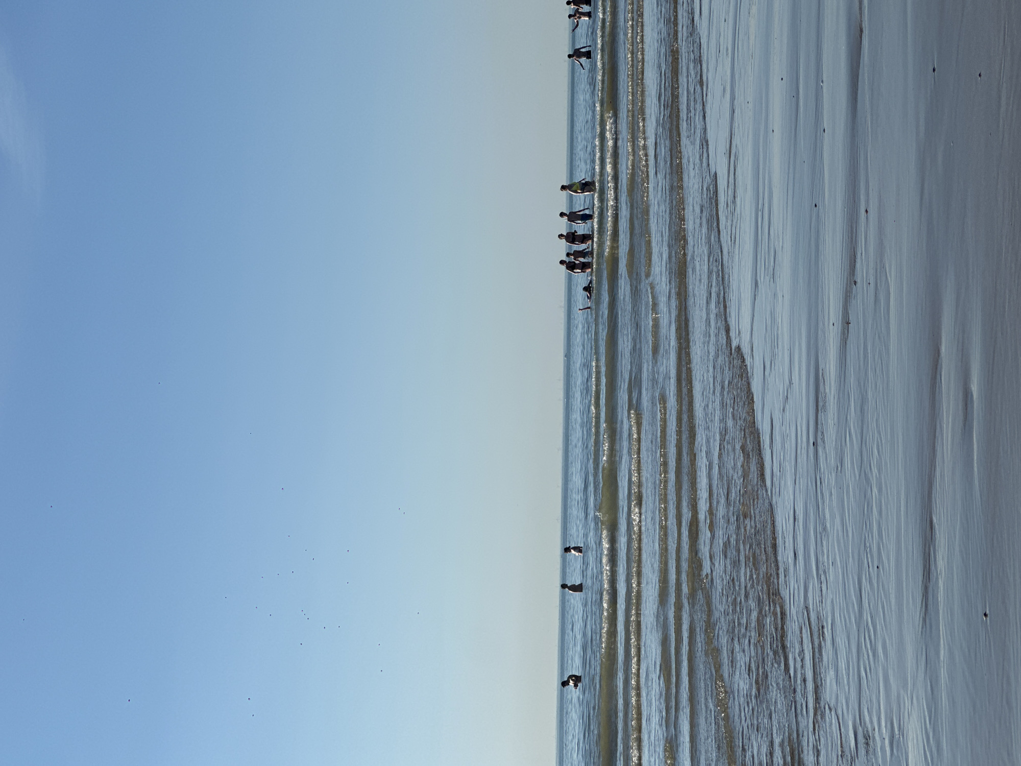 The image depicts a sunny day at the beach in Zandvoort, Netherlands, with a clear blue sky and calm waters. Several people are visible in the sea, some standing in the shallow water and others swimming. The foreground shows the wet sand of the beach with ripples from the receding waves. In the distance, the horizon is lined with a faint outline of what could be buildings or structures, possibly indicating the proximity of the town. The overall scene conveys a relaxed and leisurely atmosphere of a day at the seaside.