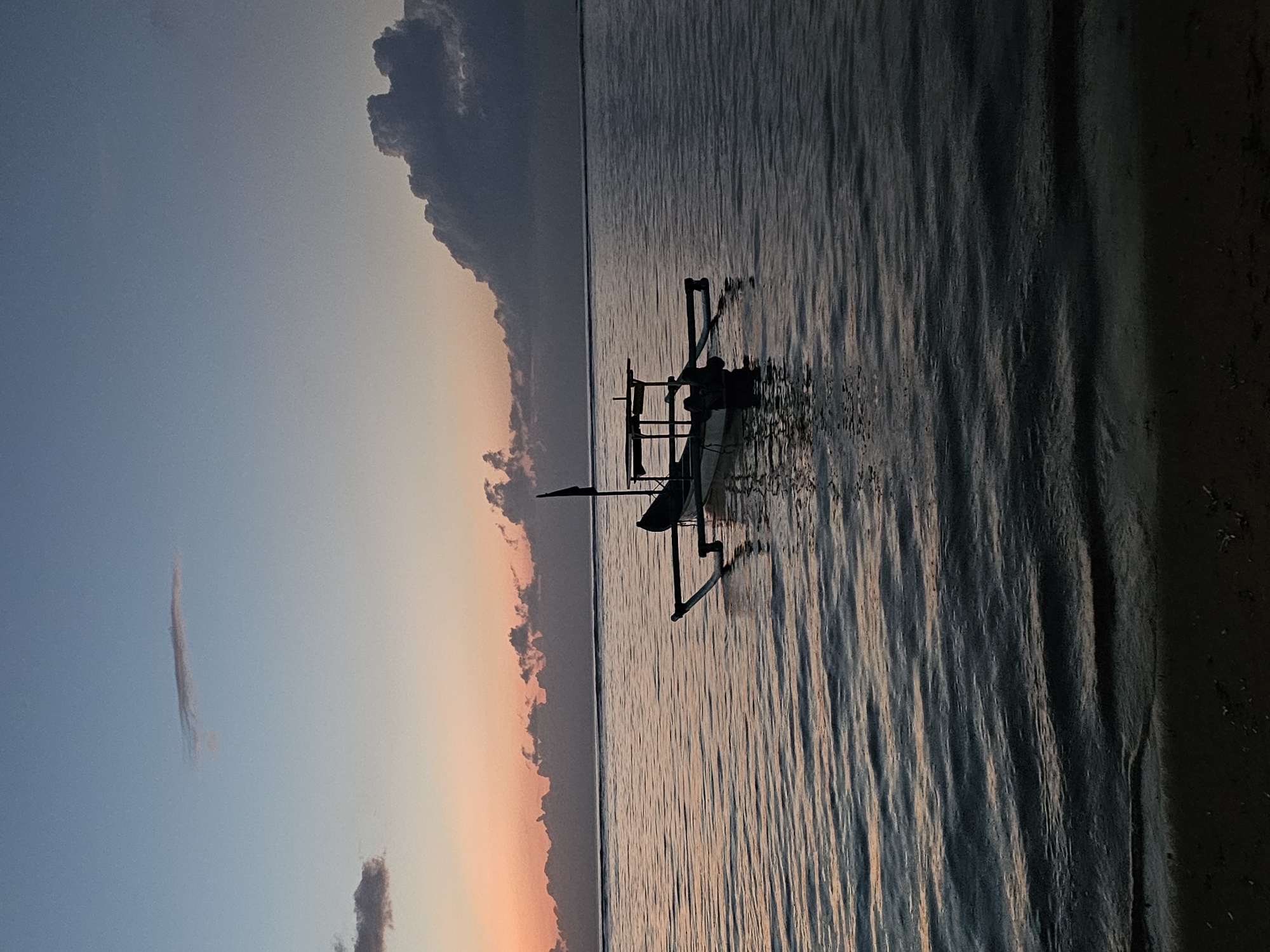 The image depicts a serene ocean scene at dusk, likely in Denpasar, Indonesia, given the presence of a traditional Balinese fishing boat. The sky transitions from a pale orange and pink near the horizon, indicative of a sunset, to a deep blue overhead. Scattered clouds are visible, with larger, darker formations on the right side of the frame. The sea is calm, with gentle ripples reflecting the colors of the sky. A lone outrigger canoe, a common sight in Indonesian waters, is anchored in the middle distance. It is empty and appears to be a fishing vessel. The foreground shows a dark, sandy shoreline. The overall atmosphere is peaceful and tranquil, capturing the end of a day.