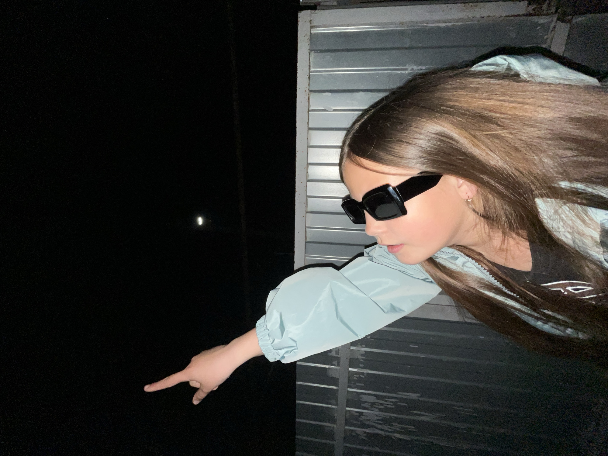 A young woman is partially visible through an opening, possibly a window or doorway, in what appears to be a metal garage door or structure. She is wearing a light blue jacket and black sunglasses. Her right arm extends outward, with her index finger pointing downward into the darkness.
The background on the left side of the image is black, with a small, faint light visible in the distance. The right side of the image shows the metal door structure, with horizontal lines indicating the individual panels of the door.
The image was taken in Novopokrovskaya, Russia.