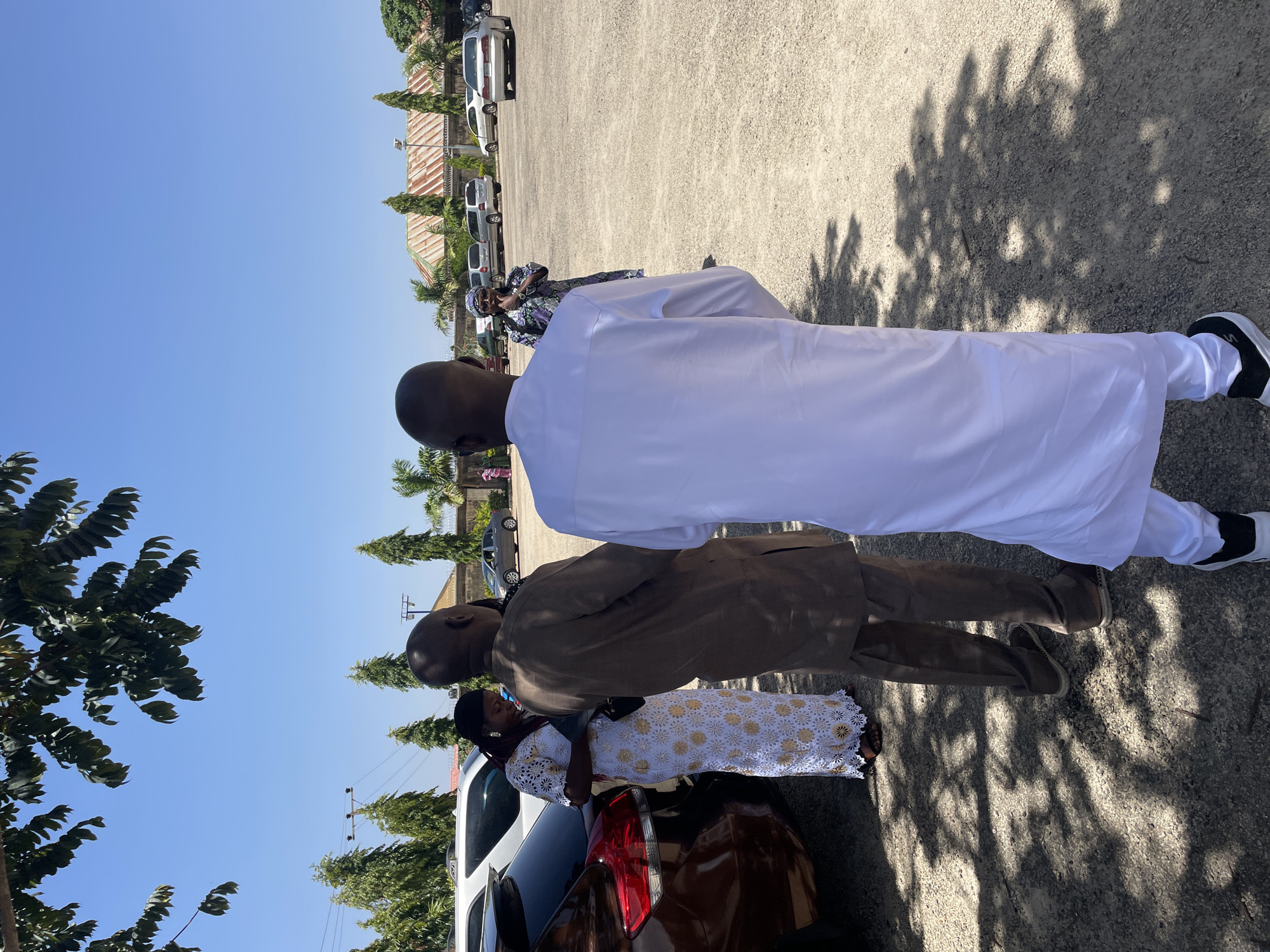 The image depicts a sunny outdoor scene in Jos, Nigeria. Three people are standing near a brown car. The person closest to the camera wears a brown suit and the person to the right wears a white robe and black and white sneakers. A woman is standing next to the car, wearing a white dress with gold patterns. Other cars are parked in the background, near a building with a corrugated roof. Trees are visible both on the left side and in the background. The sky is clear and blue. Tree shadows are cast on the ground.