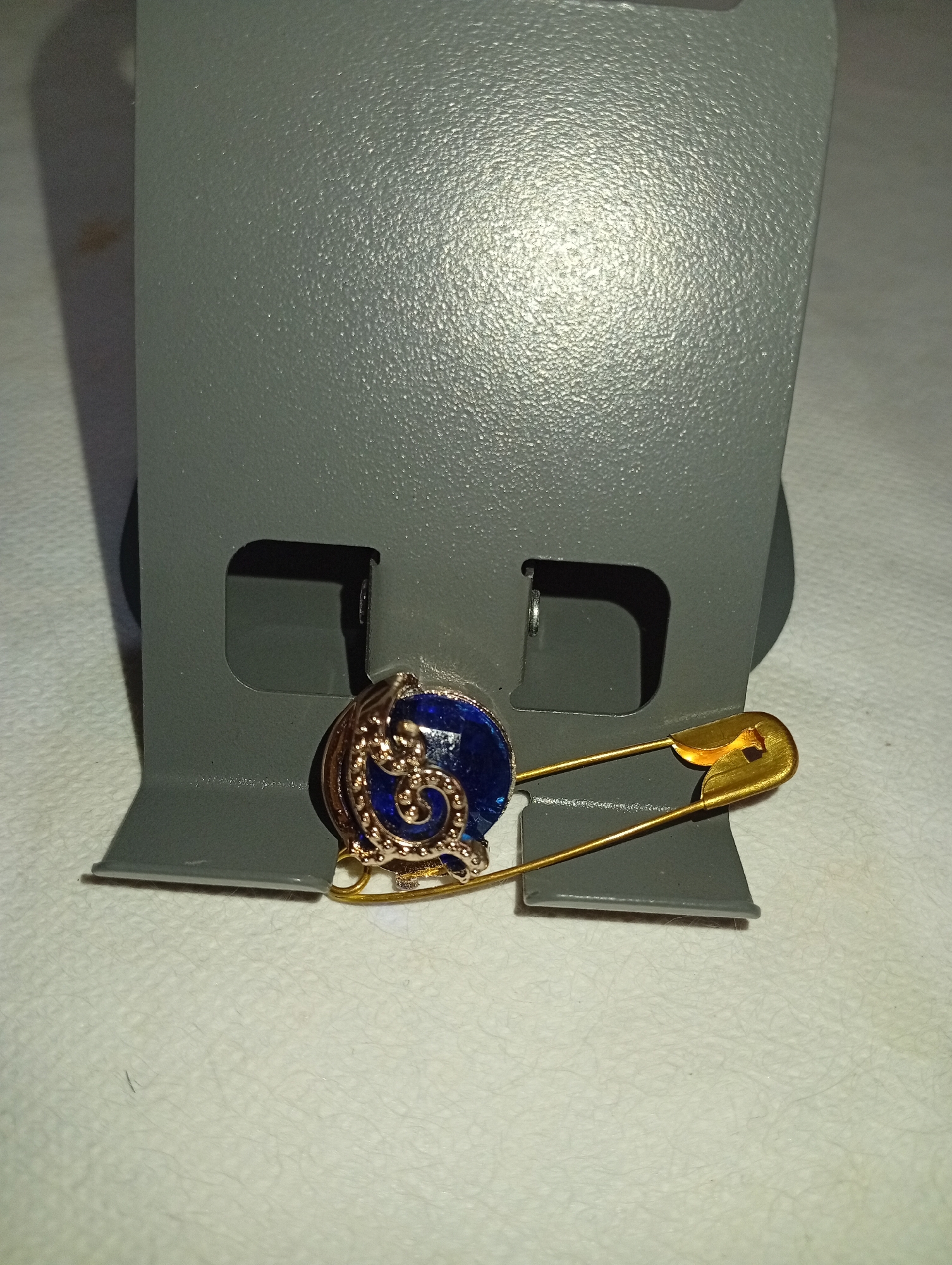 A close-up, eye-level shot shows a brooch with a blue stone and gold filigree, attached to a golden safety pin. The brooch is pinned to a gray display stand, which has two square cutouts. The stand is placed on a textured white surface. The background is out of focus, with a shadow cast by the stand on the left side. The image was captured in Seýdi, Turkmenistan.