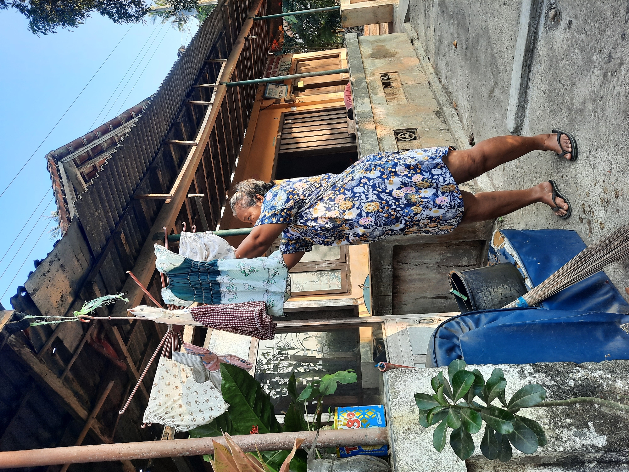 An elderly Indonesian woman with graying hair pulled back, wearing a blue floral dress and black flip-flops, is hanging laundry on a drying rack outdoors in front of a building. She is positioned on the right side of the frame, reaching up to attach a garment. The drying rack is a metal structure with multiple arms, holding various pieces of clothing, including a patterned dress, a checkered cloth, and infant clothing.
The background shows a rustic building with a corrugated roof and wooden supports, suggesting an outdoor covered area. A window and a doorway are visible on the building's facade. To the left of the woman, there is a concrete ledge with a plant in a pot, and a blue container, a black bucket, and a broom are also visible. The ground is concrete paving. The scene is bathed in natural light, indicative of daytime. The location is identified as Andong, Indonesia.