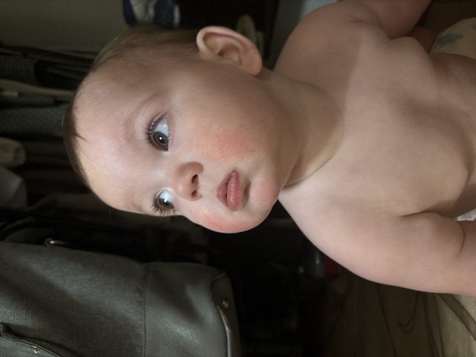A baby with fair skin and light brown hair is looking at the camera. The baby has a slightly flushed face and dark eyes with long eyelashes. The baby's lips are full and pink. The upper body of the baby is visible, and it appears to be lying on its back. A gray bag is partially visible in the background.